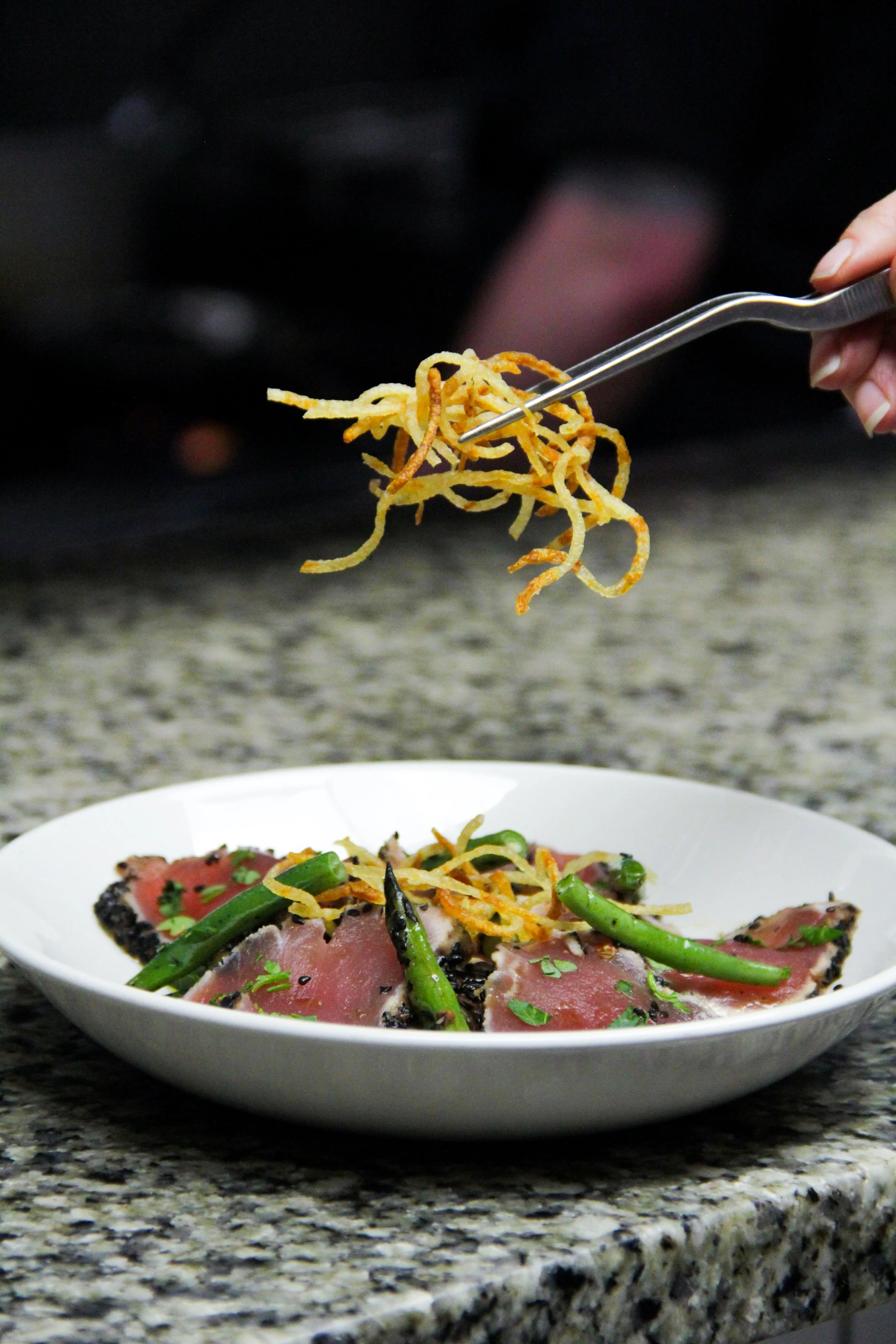 Close-up of a hand holding a fork with crispy fried noodles over a plate of seared tuna with green beans, garnished with chopped herbs on a granite countertop.
