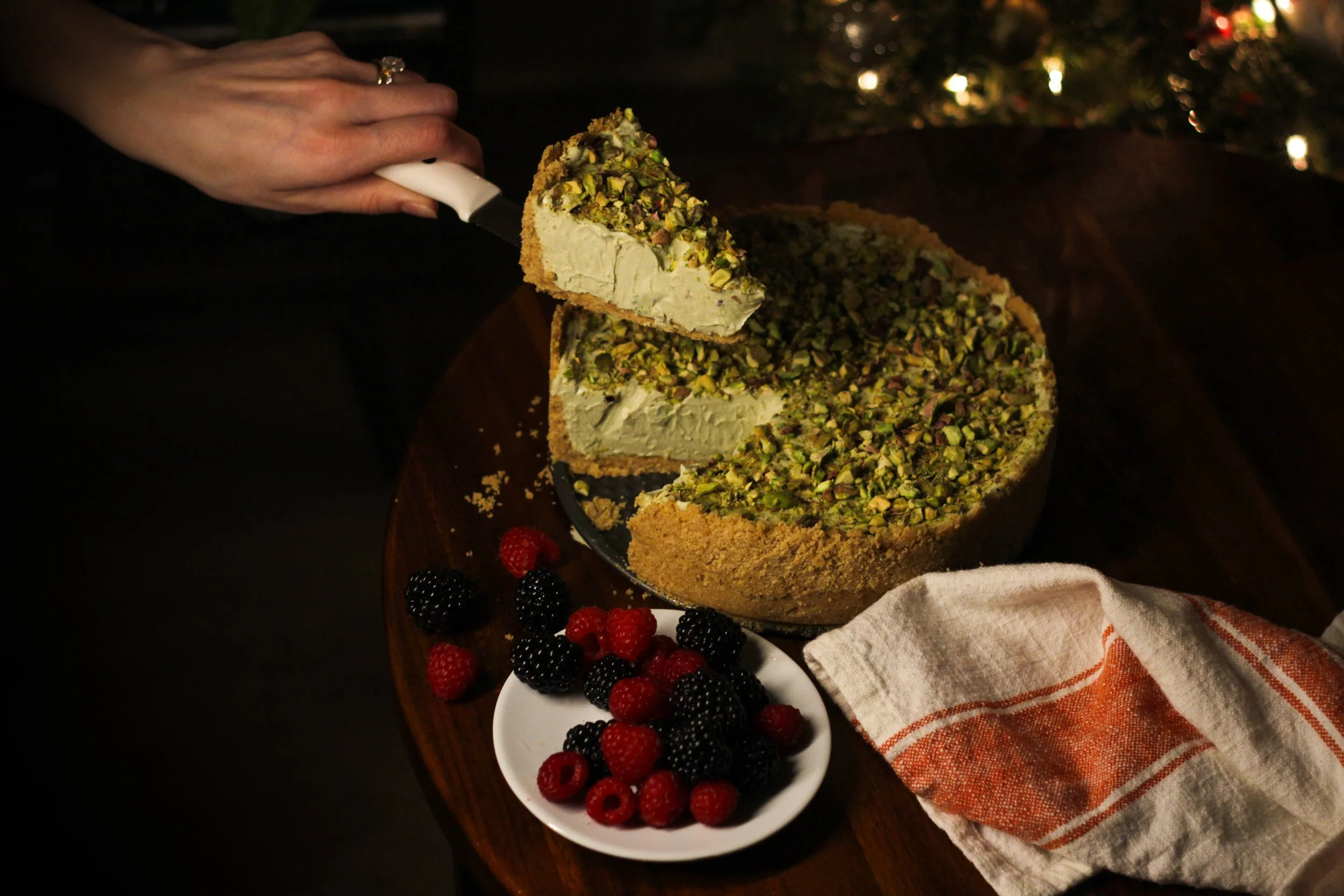 A person is cutting a cheesecake topped with chopped pistachios, with a plate of fresh berries nearby.