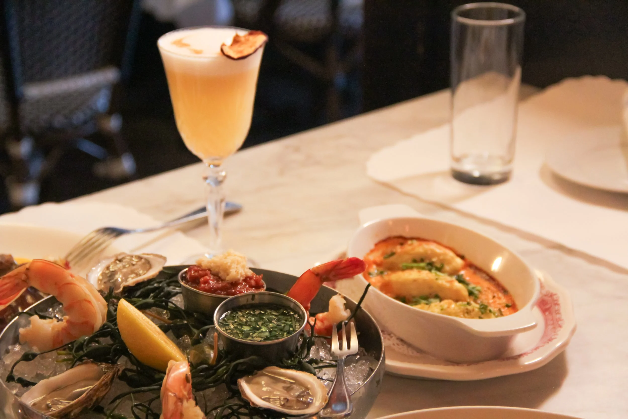 Seafood dish on ice with shrimp, oysters, and lemon wedge, served with two dipping sauces, paired with a glass of orange cocktail and an empty glass on a white marble table.