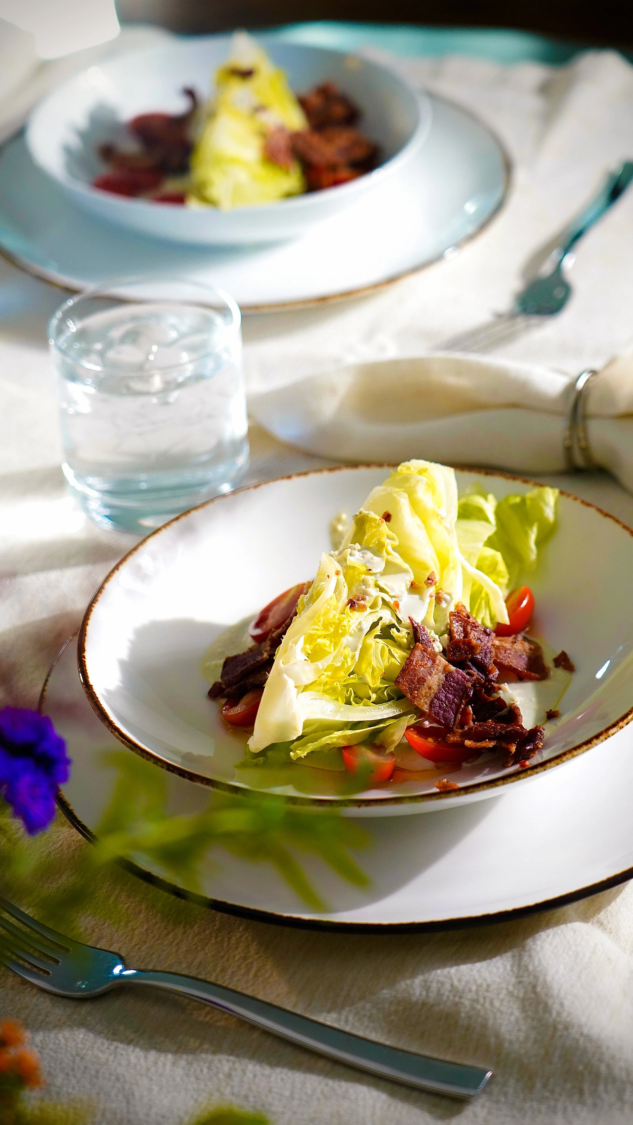 Close-up of a plated salad with lettuce, cherry tomatoes, bacon bits, and a slice of potato, on a white plate with a gold rim, set on a table with a glass of water, a fork, and a napkin in the background.