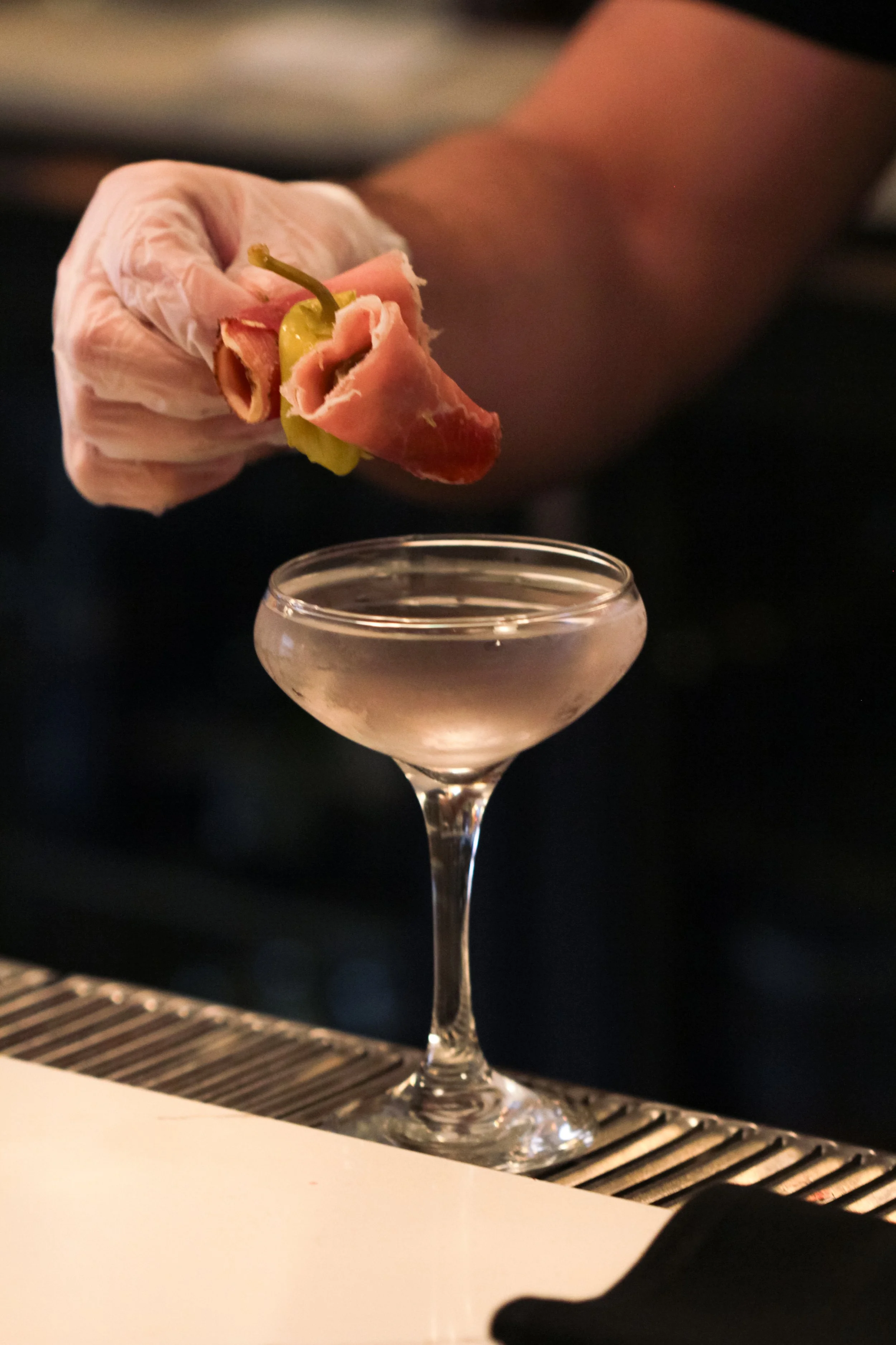 A person wearing a white glove holds a shrimp garnished with a slice of ham and a green pepper over a pink cocktail in a coupe glass on a bar counter.