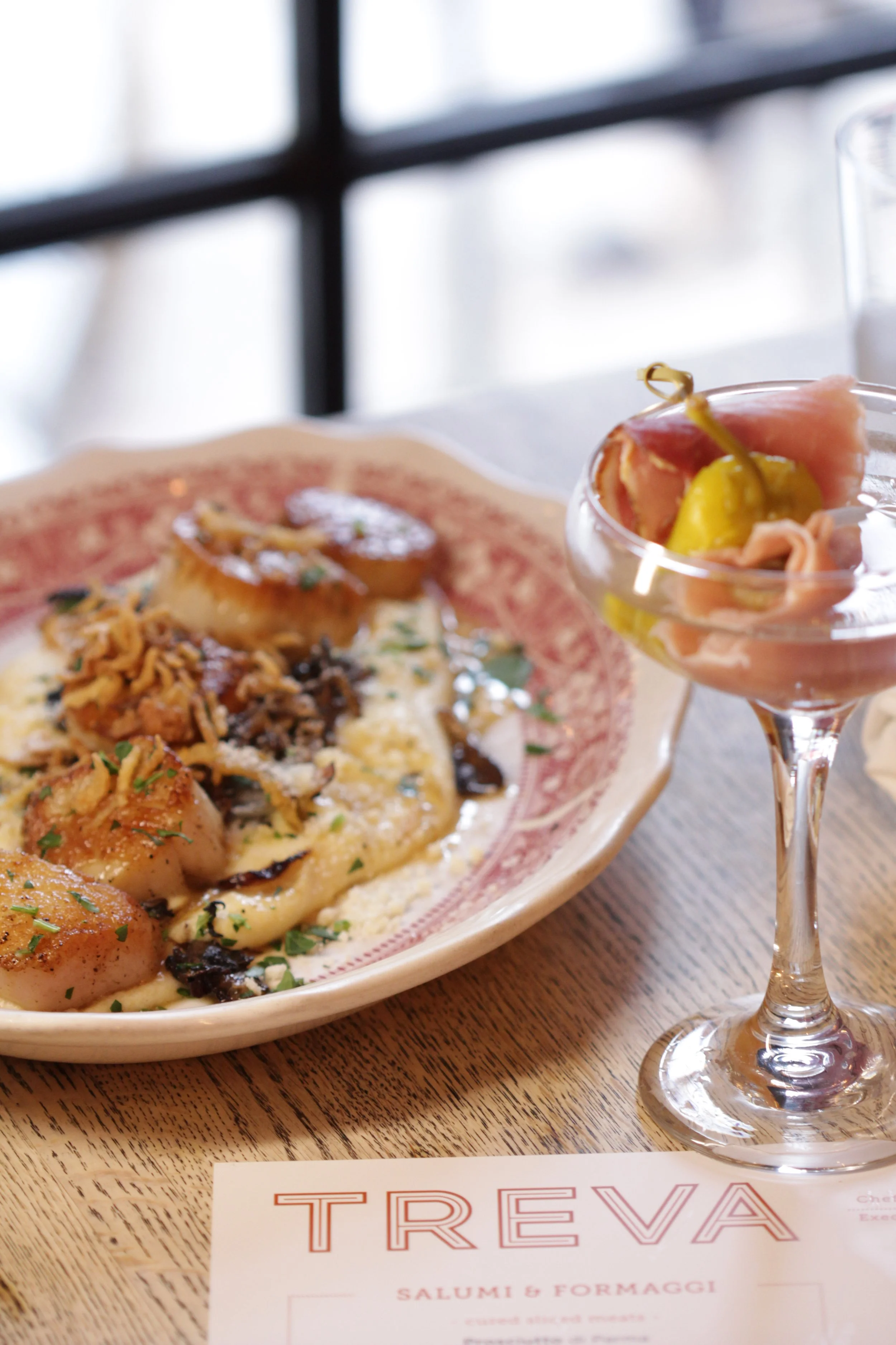 A plate of pasta with scallops, garnished with herbs, alongside a glass of cold pink drink with prosciutto, cheese, and pickles.