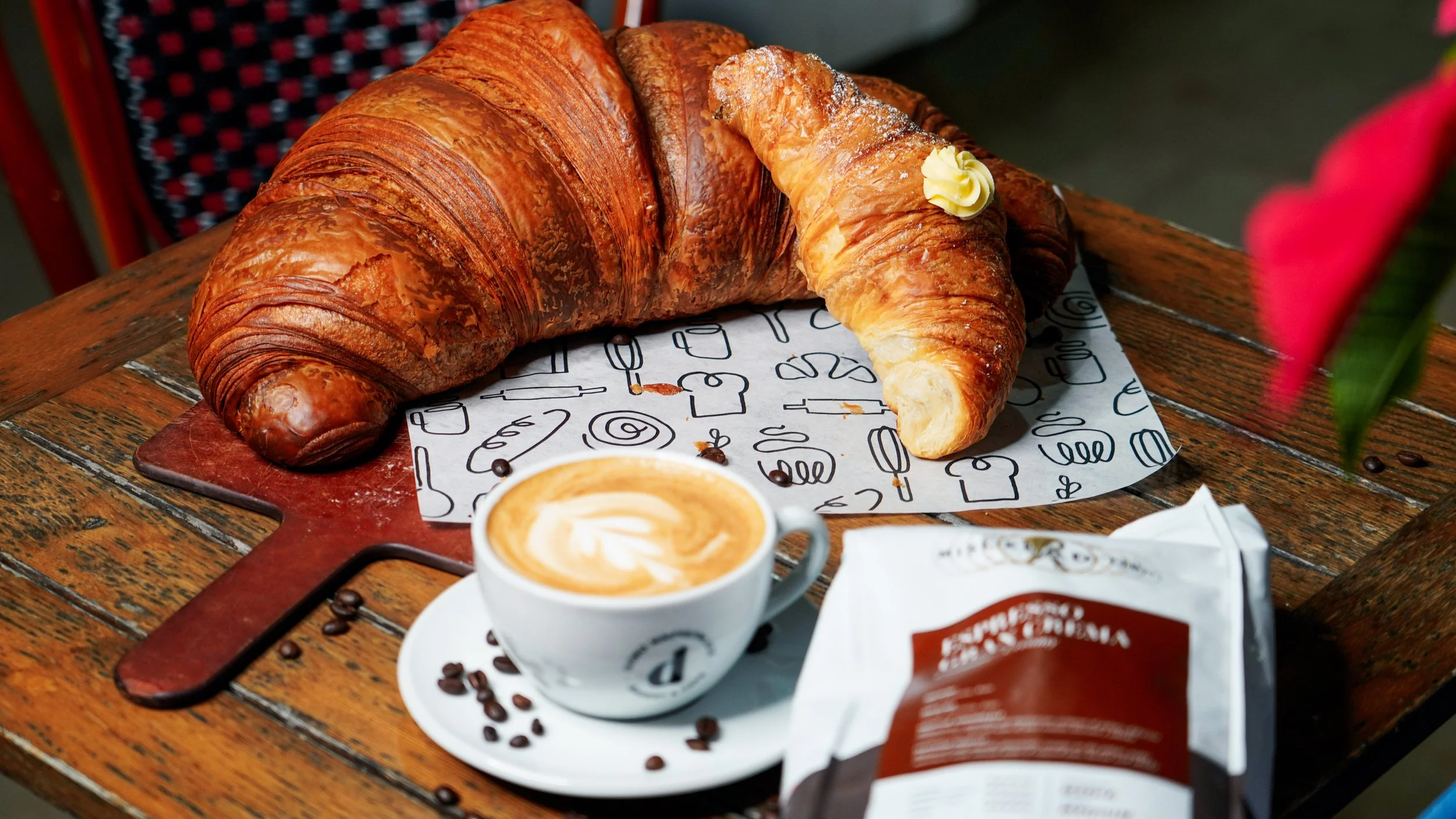 A wooden table with a croissant, a cup of latte with latte art, and a packet of coffee, with a flower in the foreground.