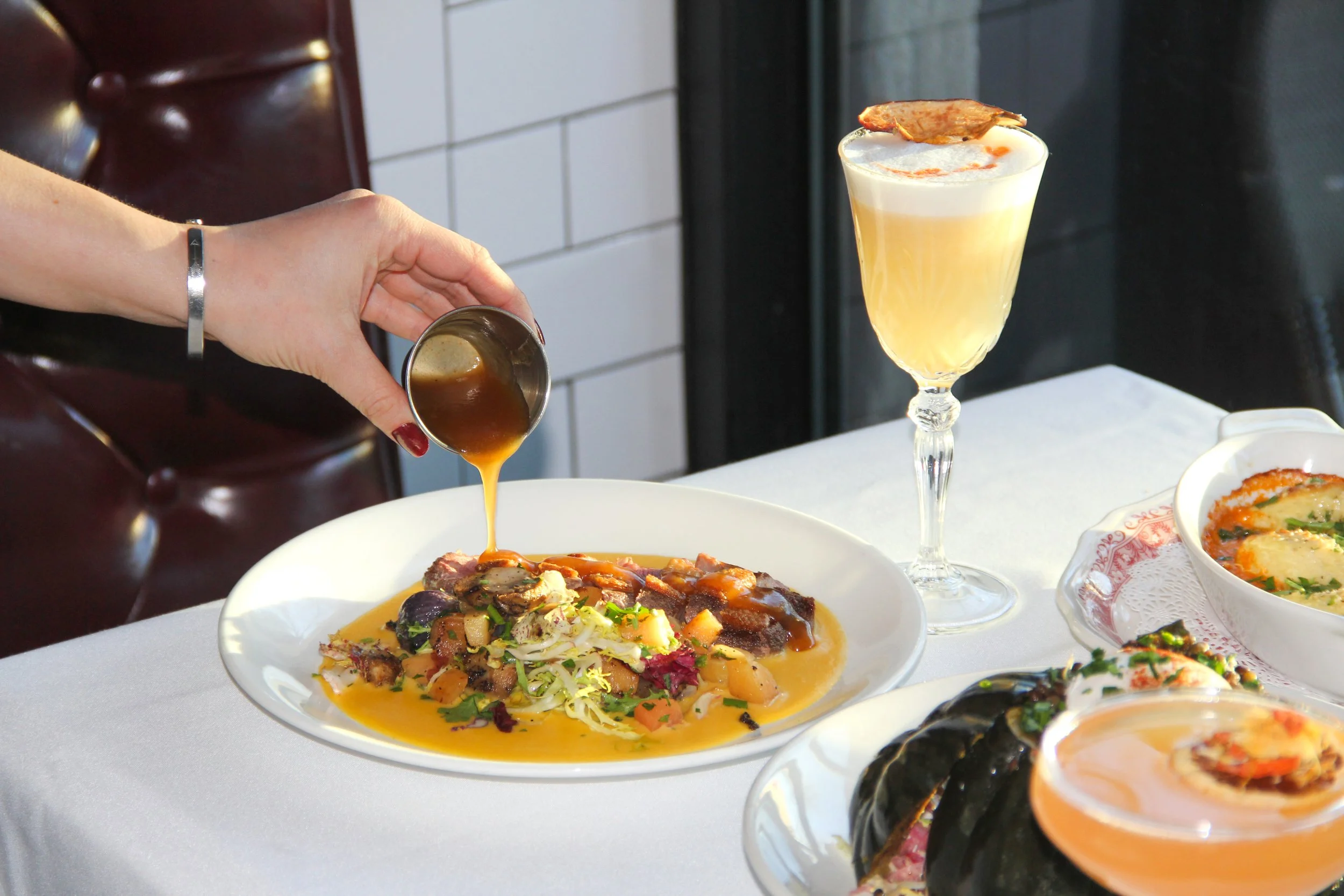 A hand pouring gravy over a plate of food, with a side of a creamy cocktail and other dishes on a white tablecloth.
