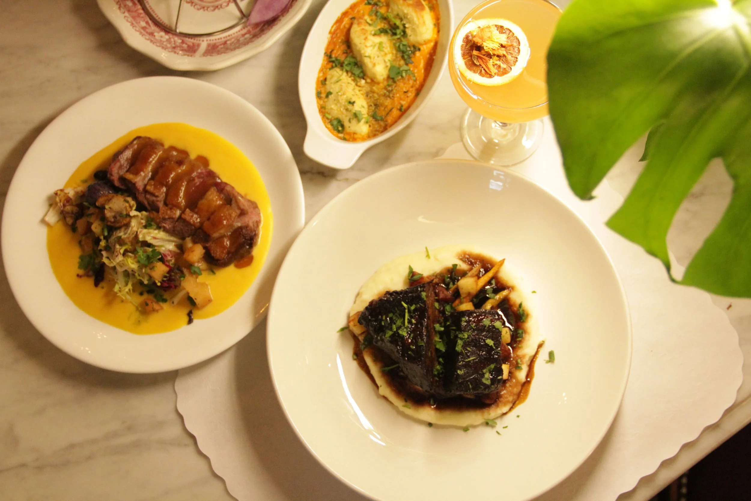 Assorted dishes including a plate with beef and vegetables in sauce, a dish with baked eggs and greens, a glass of grapefruit cocktail with lemon slice, and a side salad with greens on a marble table.