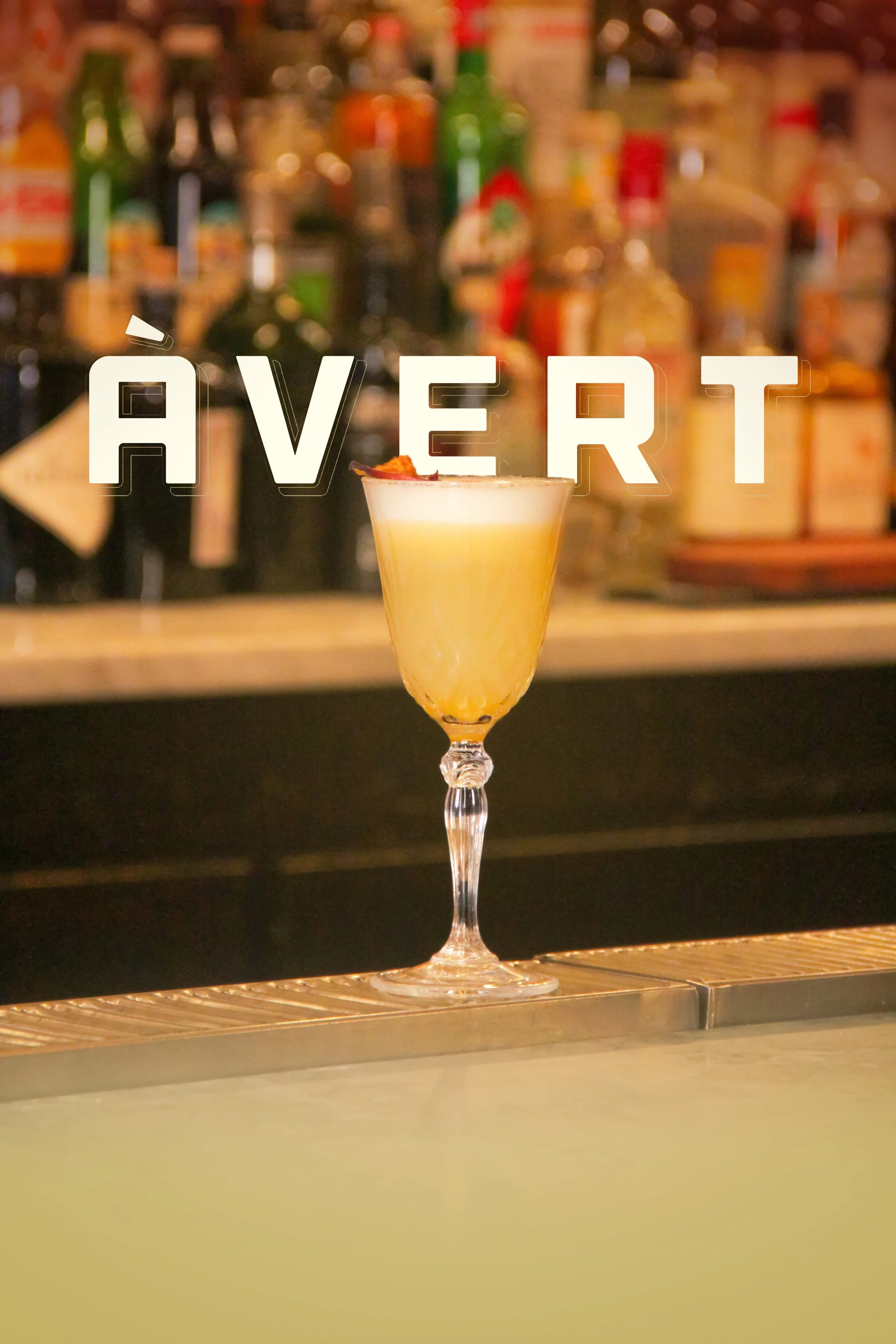 A cocktail in a tall, elegant glass with a foamy top, placed on a bar counter with a blurry background of various bottles of alcohol.