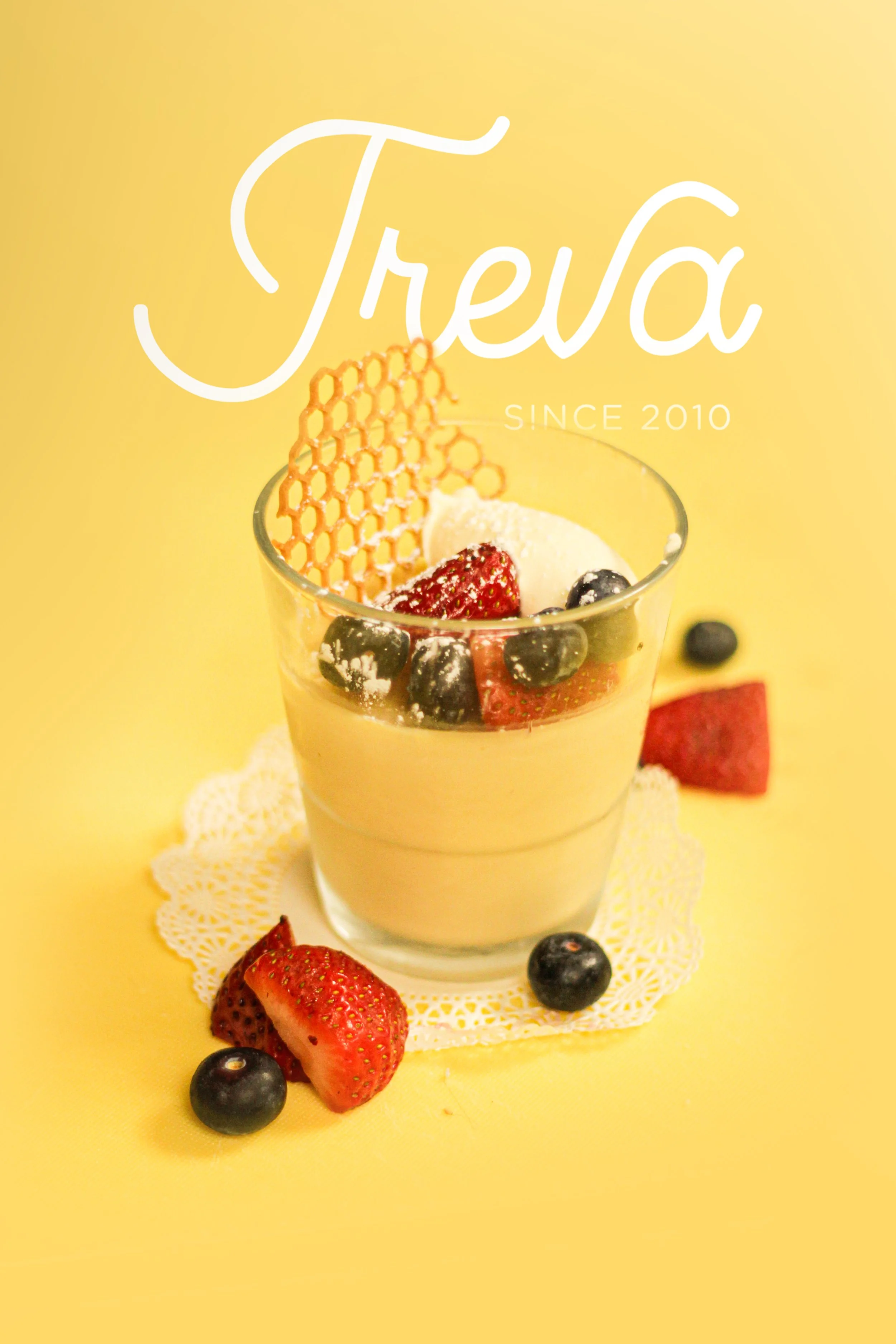 Glass of creamy dessert topped with strawberries, blueberries, and a decorative honeycomb piece on a white doily on a yellow background.