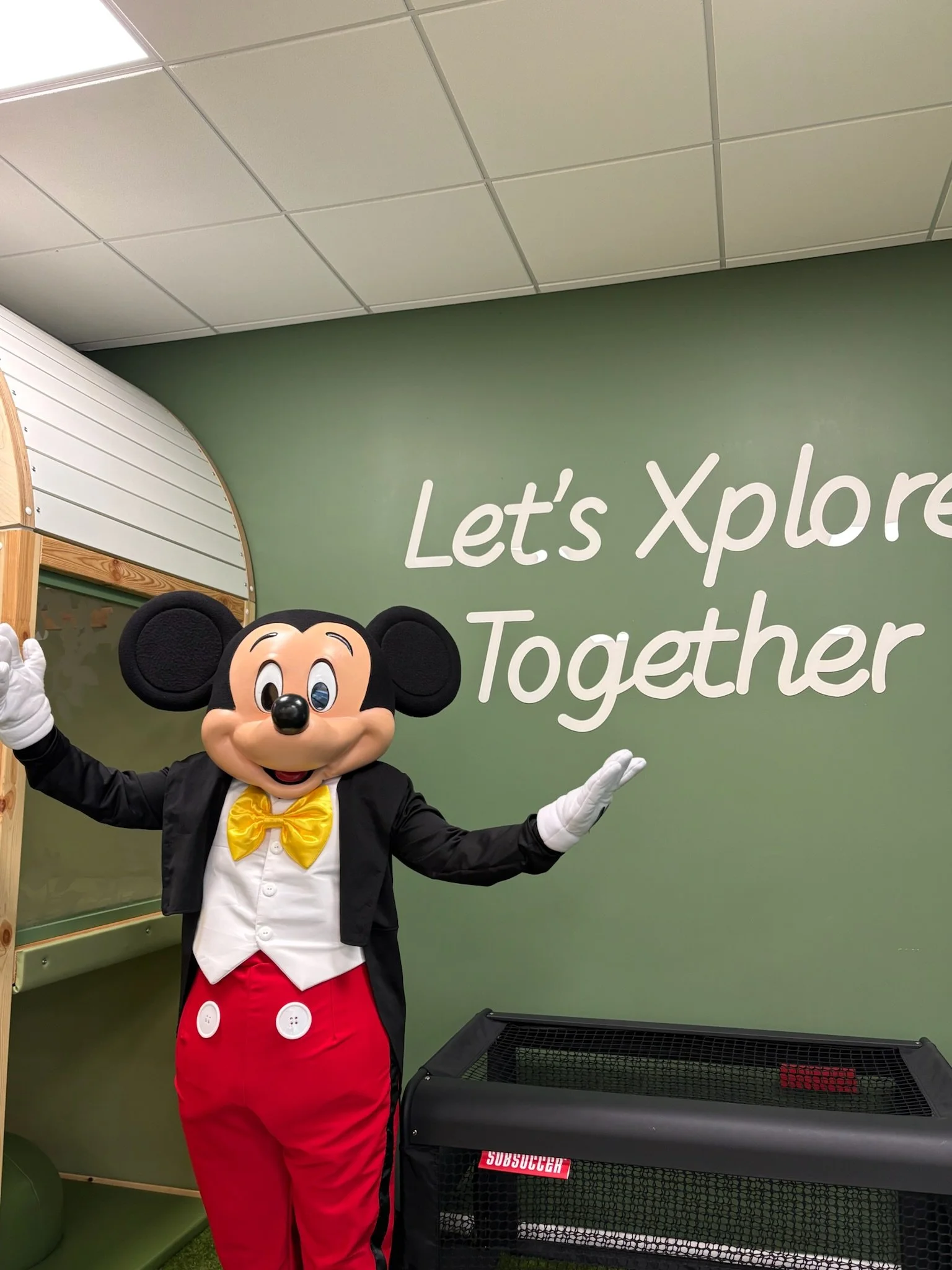 Person in Mickey Mouse costume standing in front of a green wall that says "Let's Explore Together," with a black basket labeled "Subsoccer" in the foreground.