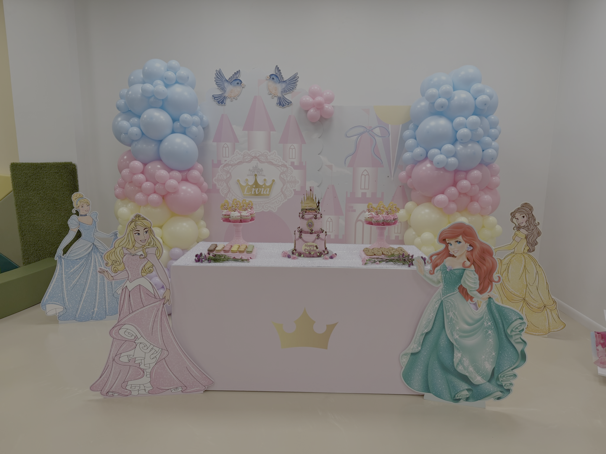 Princess-themed birthday party setup with pastel pink, blue, and yellow balloons, princess cutouts, and a pink castle backdrop featuring the name 'Livia'.