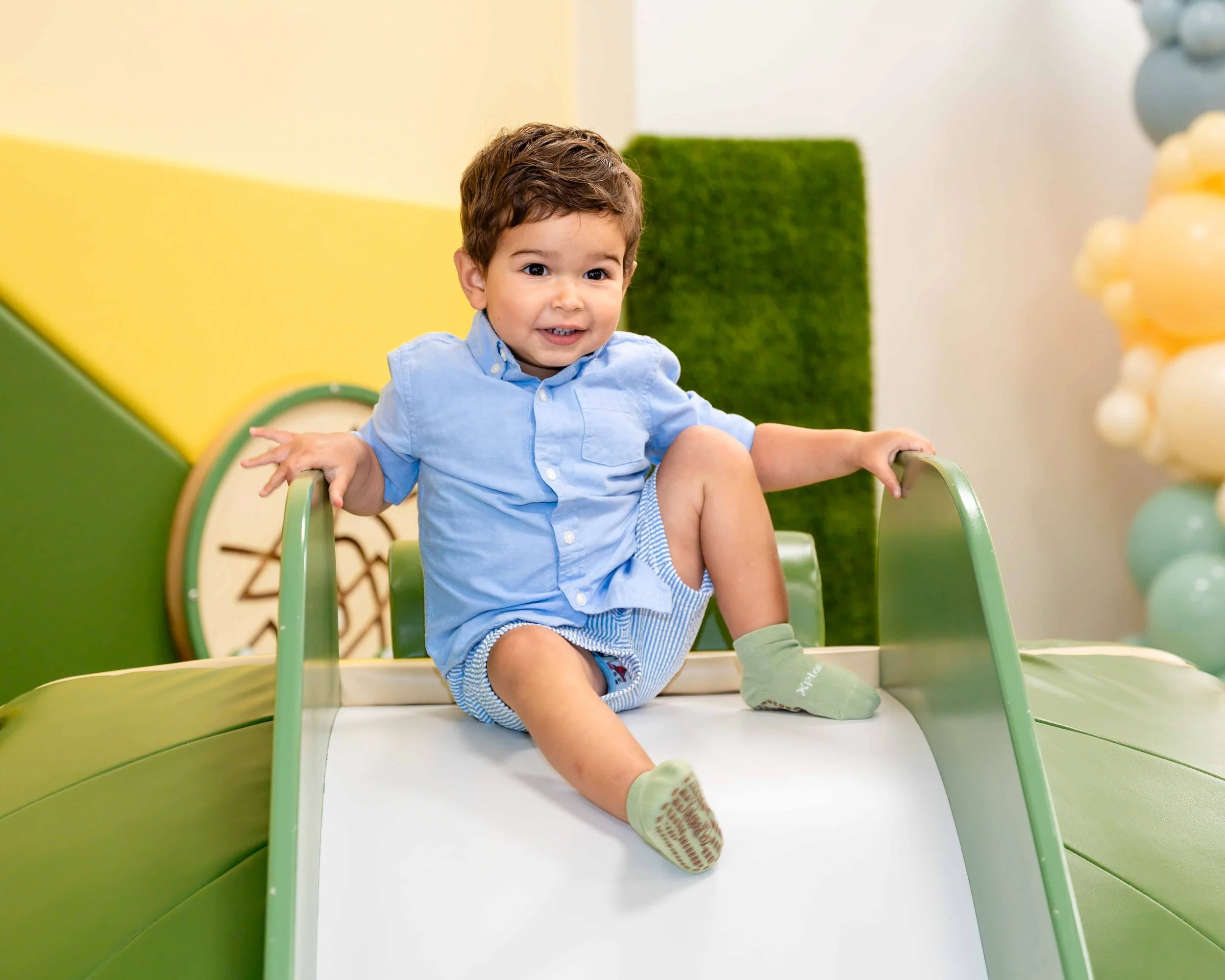 A young boy with brown hair, wearing a light blue shirt and striped shorts, sitting on a white slide at an indoor play area, smiling and holding onto the side rails.