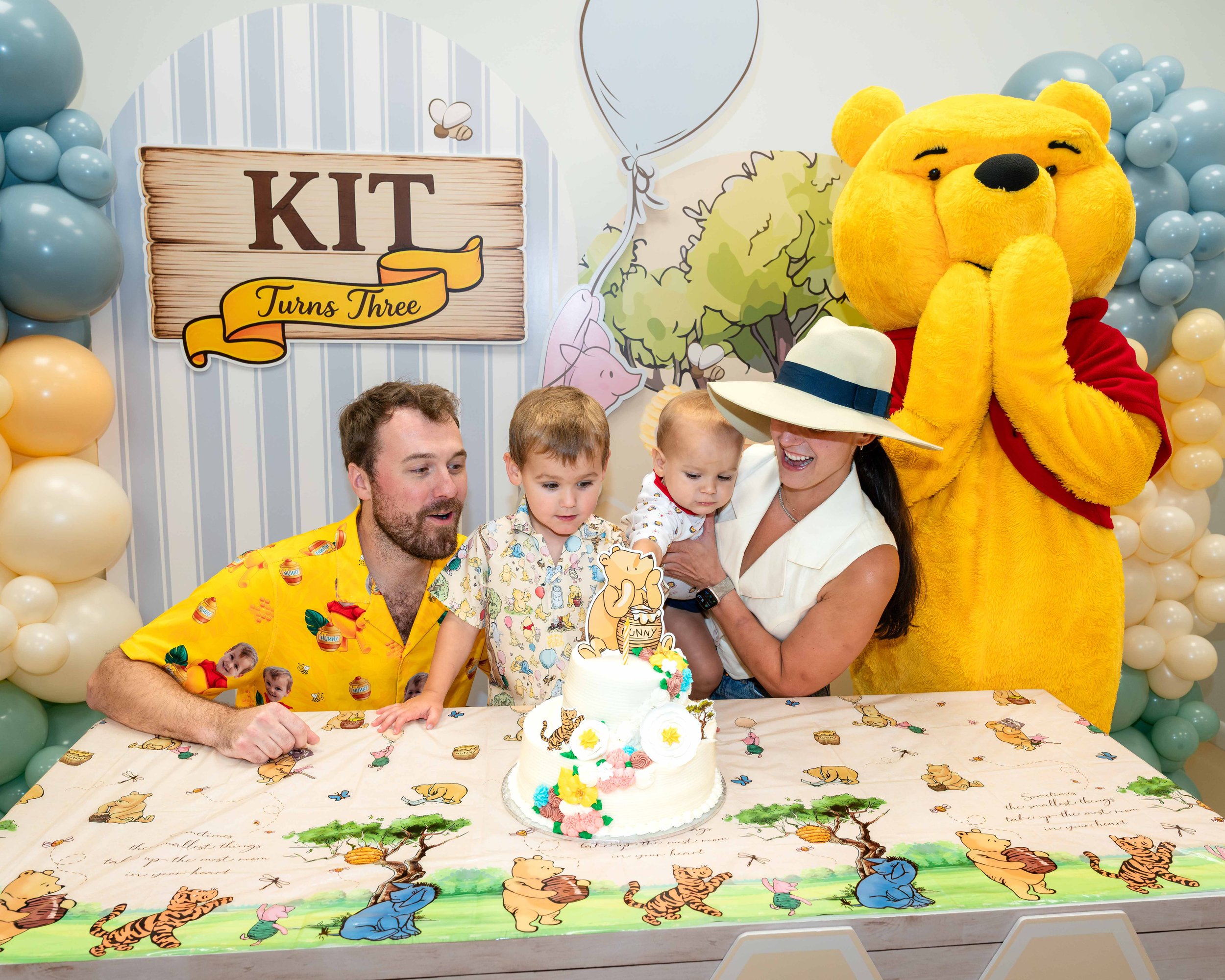 Family celebrating a child's birthday with a cake decorated with colorful flowers and animal figurines, including a tiger and an elephant. Winnie the Pooh character is present in a yellow costume, and there's a sign that reads 'KIT Turns Three' behind them, surrounded by balloons with a Winnie the Pooh and forest theme.