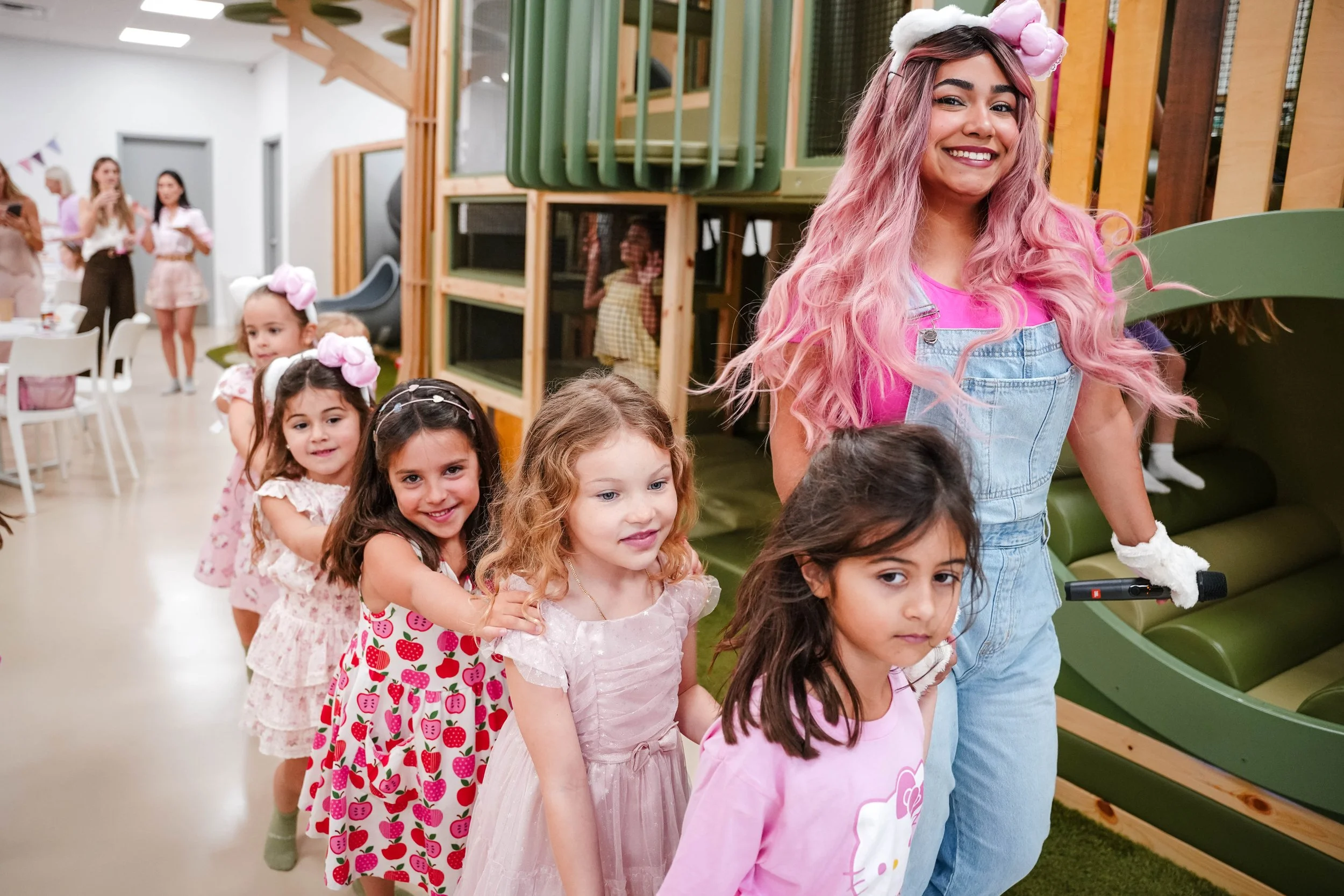 A group of young girls and a woman with pink hair and a headband with pink and white pompom, all dressed in pink and pastel-colored outfits, are standing in a line indoors. The woman at the front is smiling while holding a scooter handle, and the girls are standing behind her, with some holding onto each other. In the background, there are other women, some using their phones, and a room with modern furniture and wooden structure.
