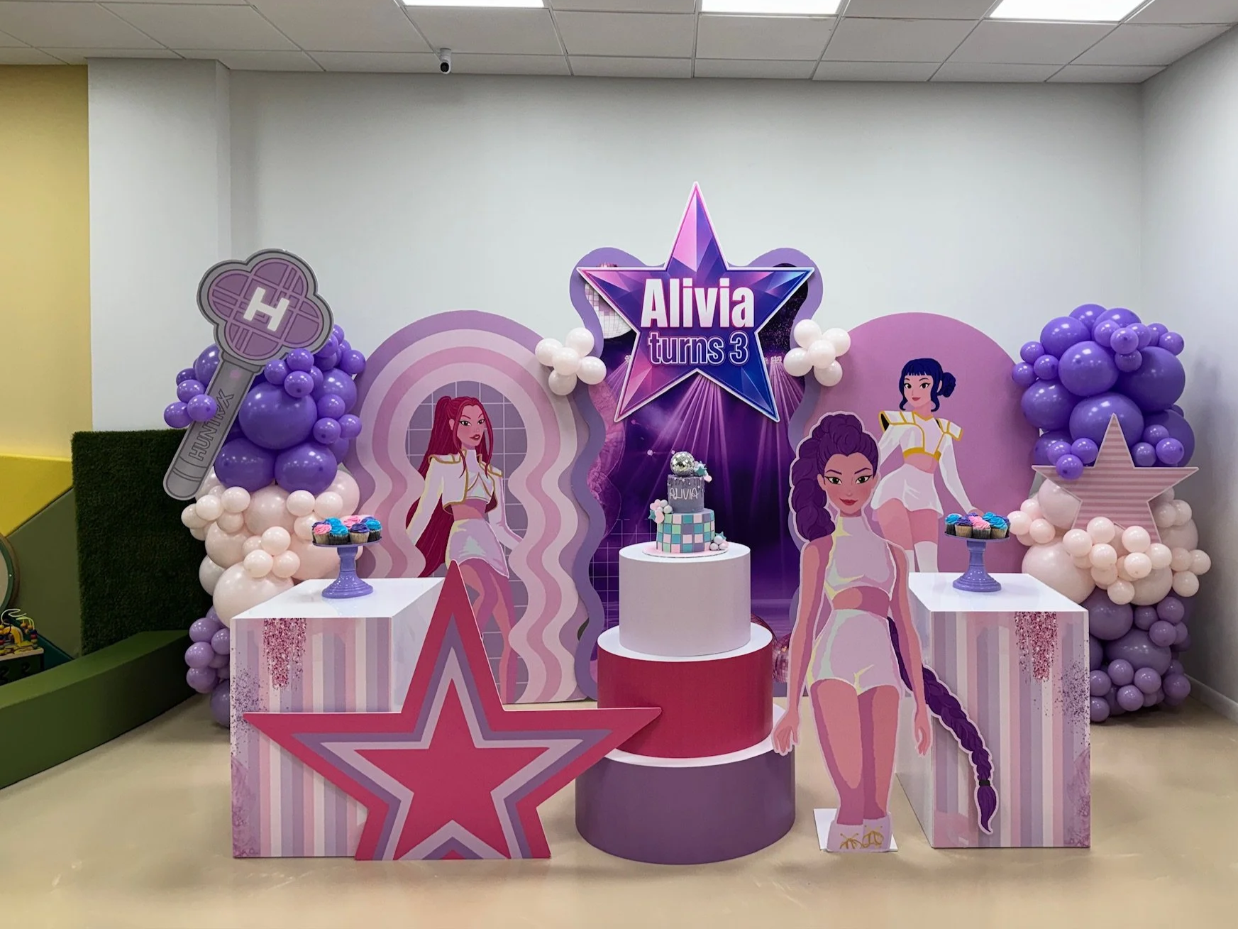 Backdrop with purple balloon decorations, cartoon characters, and a cake for a third birthday celebration. Sign reads 'Alivia turns 3'.