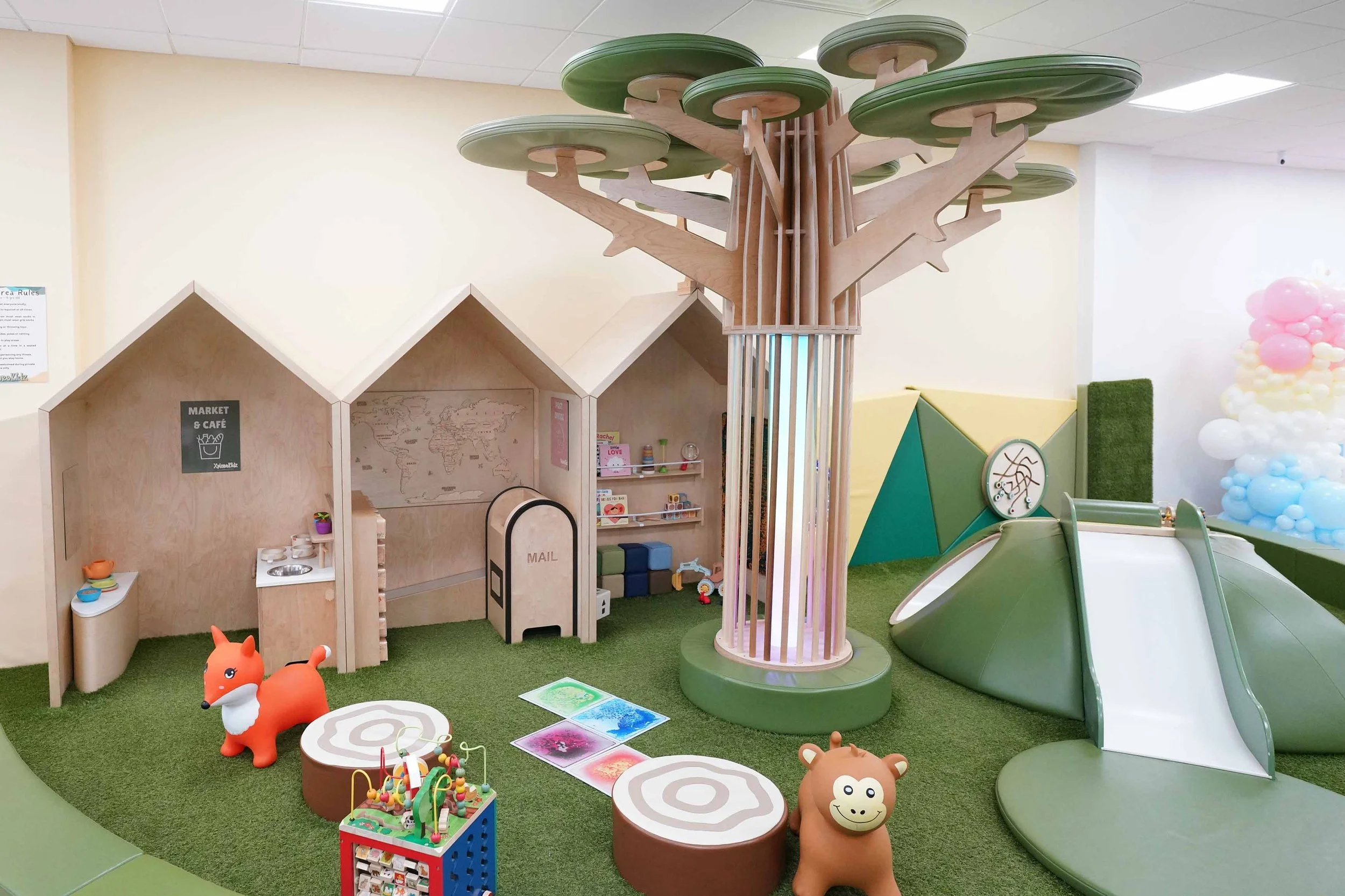 Indoor children's play area with a tree-shaped climbing structure, a small market and cafe playhouse, slides, and animal-shaped soft toys. Green carpeted flooring and colorful balloons in the background create a playful environment.