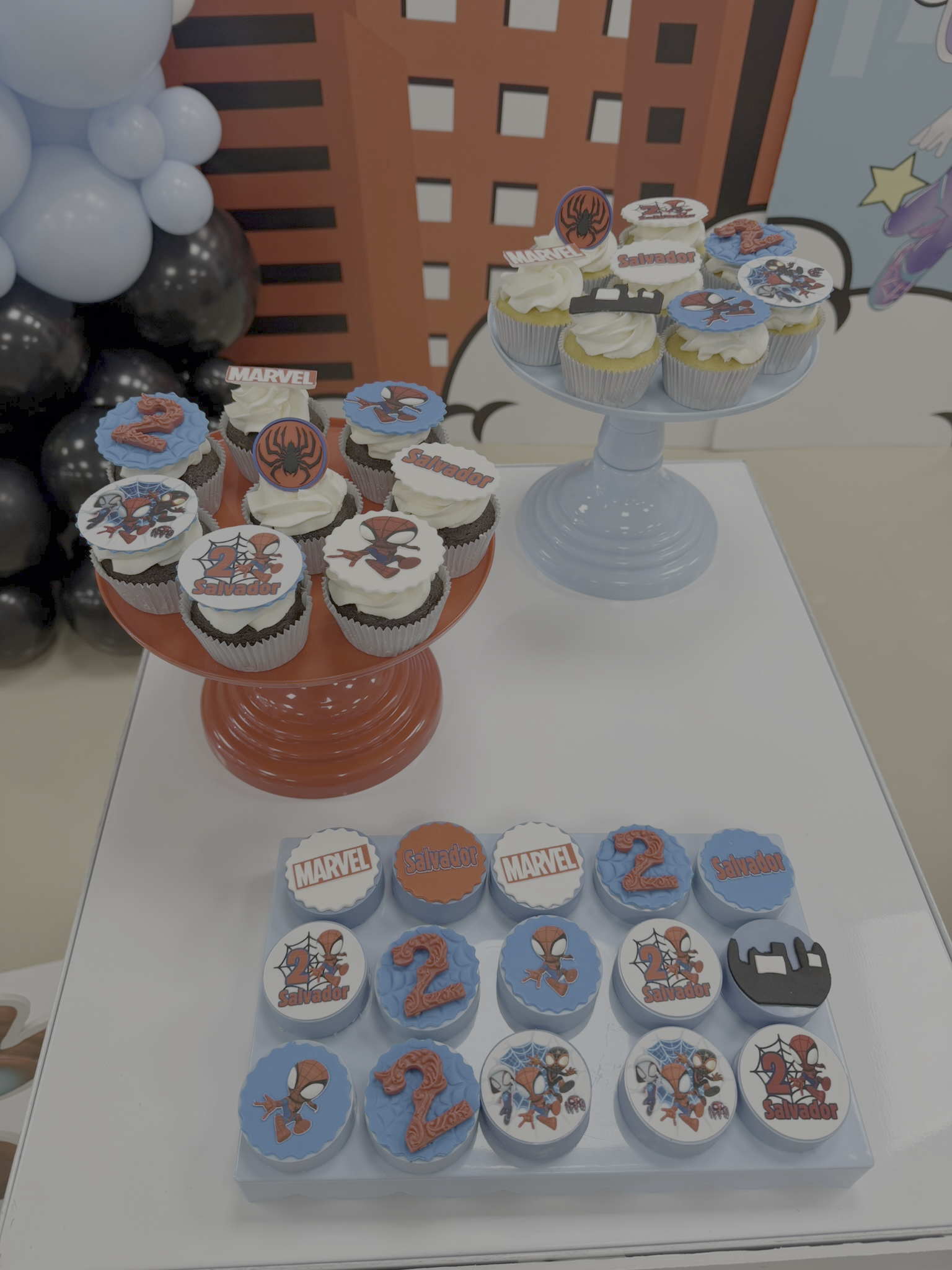 Cupcakes with Marvel Spider-Man themed decorations, some with the name Salvador, and others with the number 2 and Spider-Man images. A larger display with cupcakes on a two-tiered stand and smaller cupcakes on a tray, all decorated in blue, white, black, and orange colors.