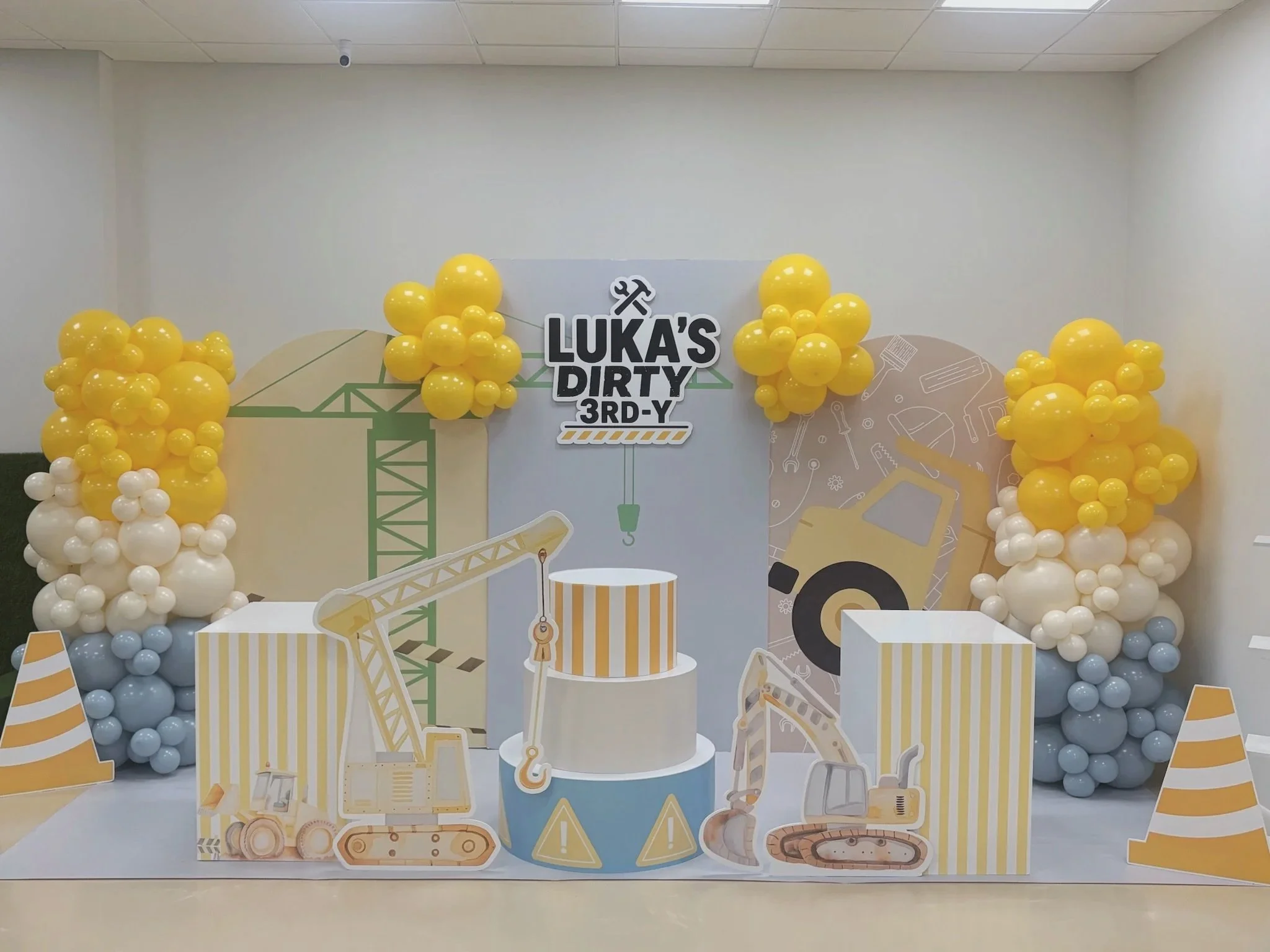 Decorative display with construction-themed cutouts and yellow and white balloons. Center sign reads "Luka's Dirty 3rd-Y."