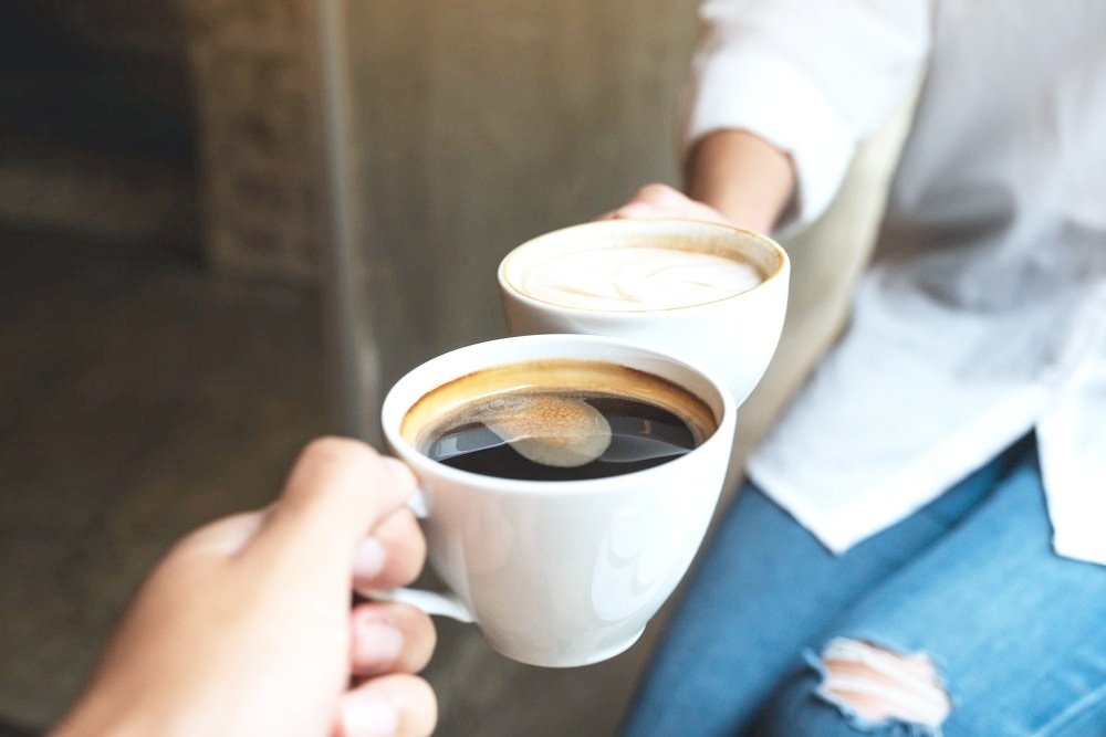 Two people holding white cups of coffee, one with foam, possibly cappuccino, and the other black coffee, indoors.