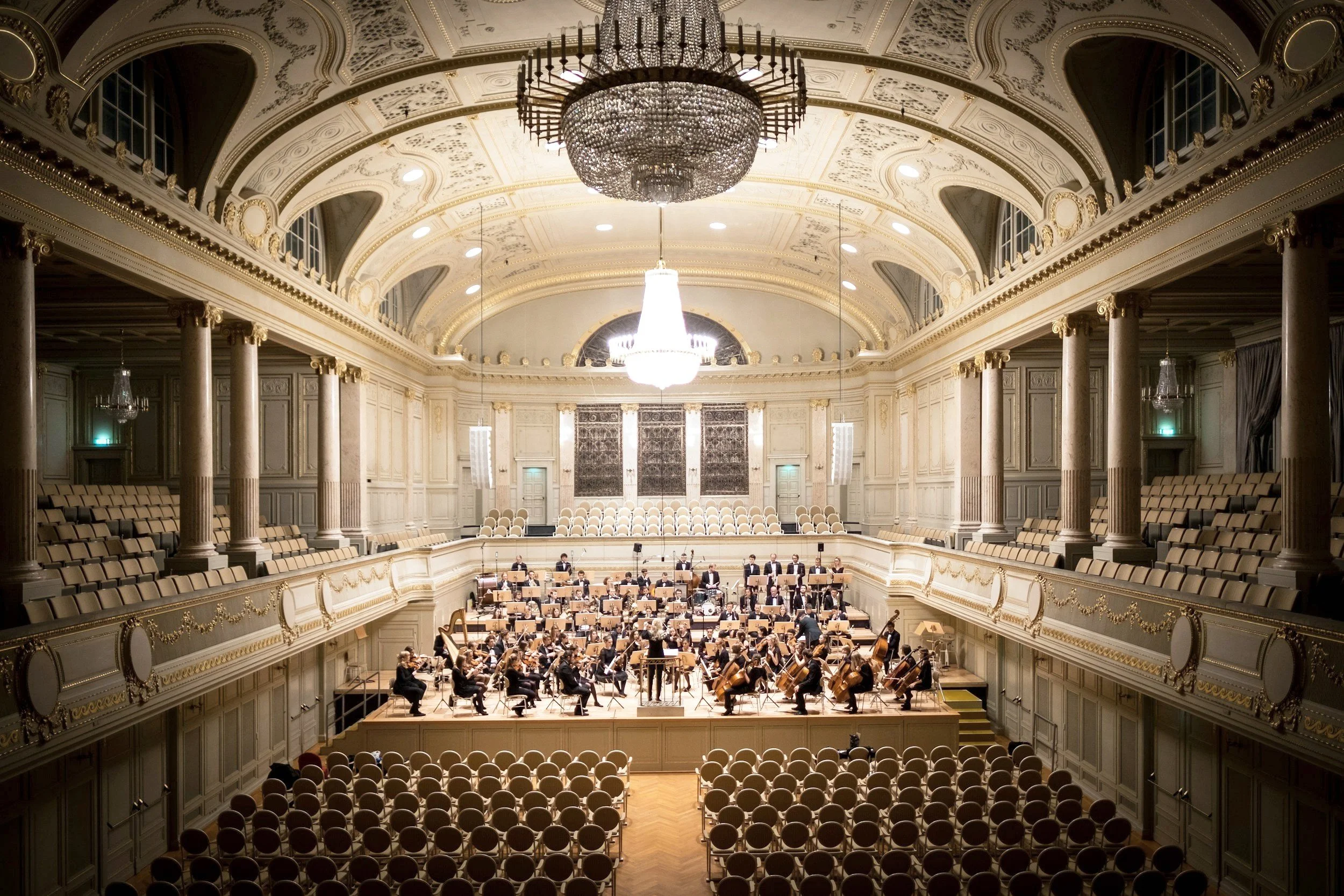 Orchestra performing in a grand concert hall with ornate architecture and chandeliers.