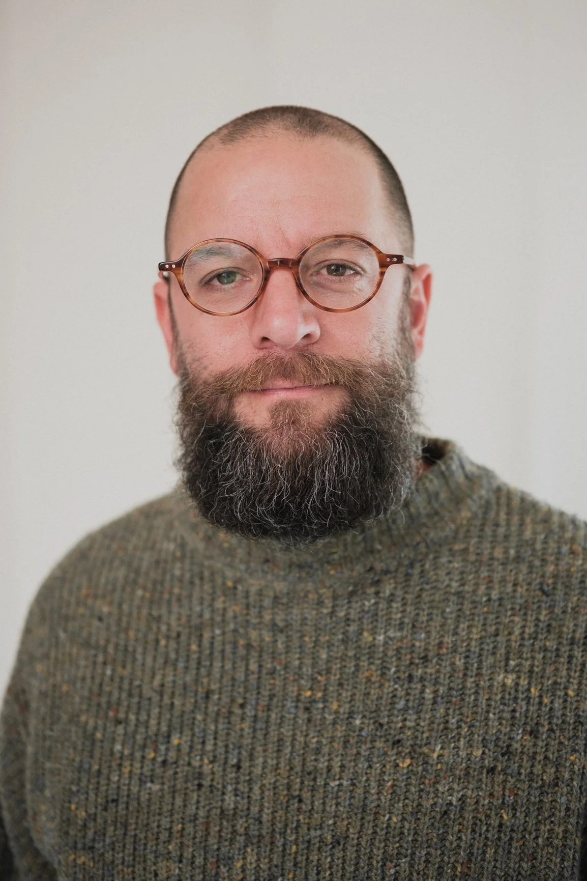 Close-up photo of a man with a short beard, bald head, glasses, and wearing a brown patterned sweater, standing against a plain light background.