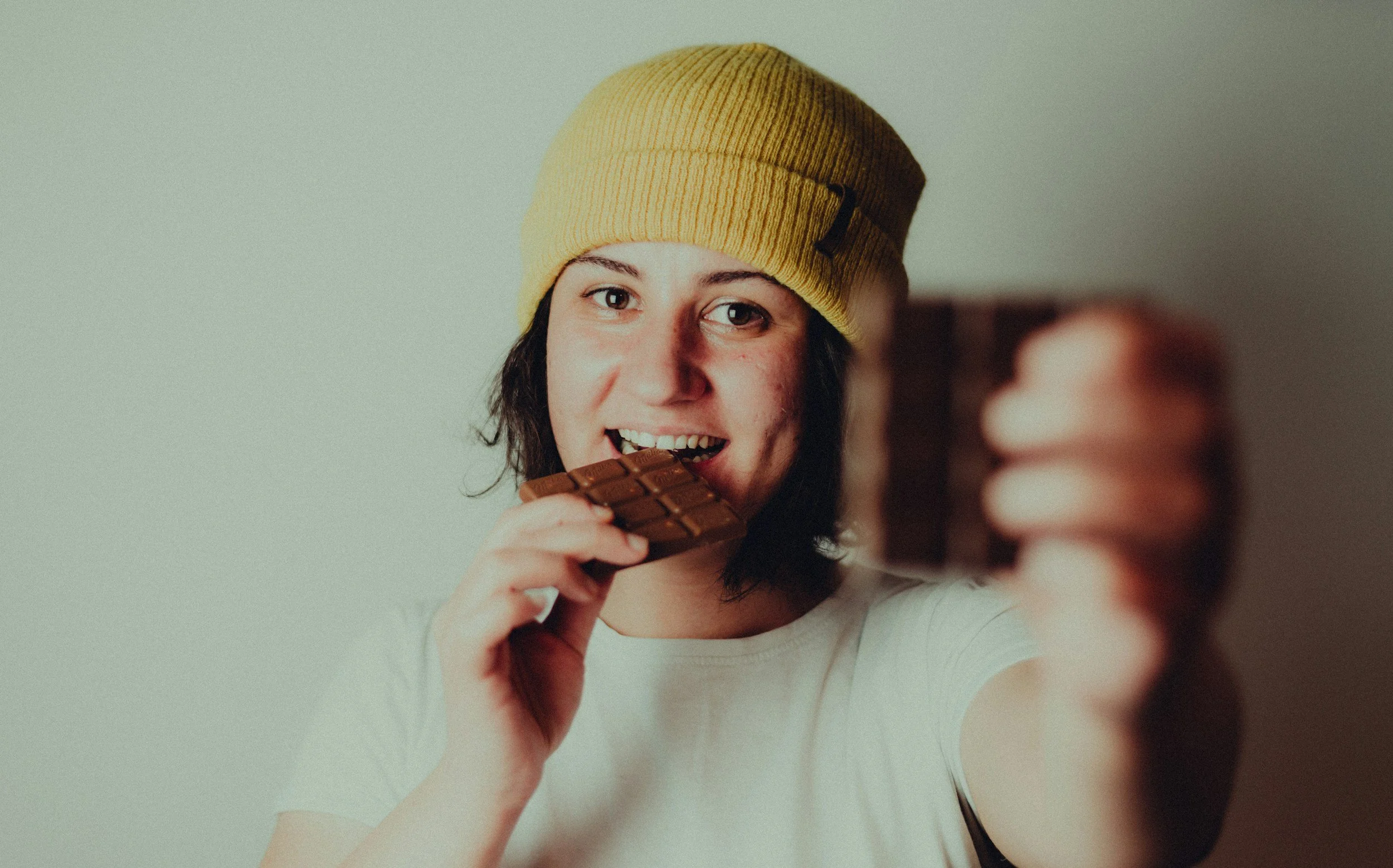 Young woman wearing a yellow beanie, taking a selfie and holding a chocolate bar close to her mouth.