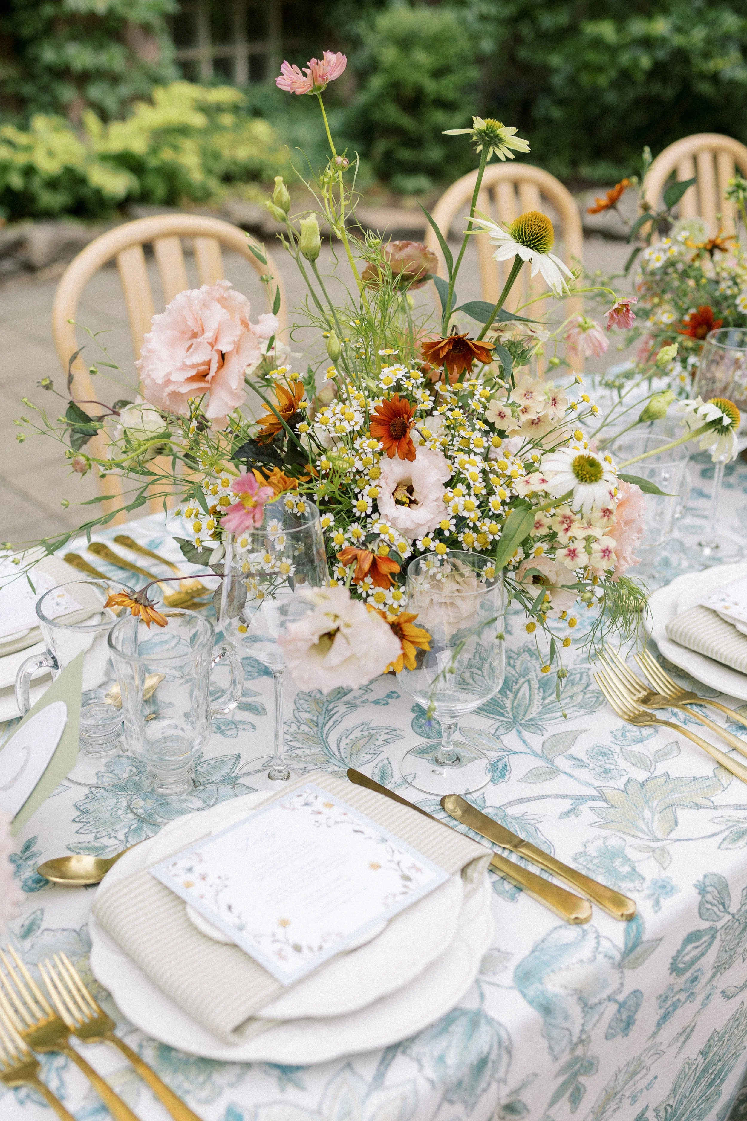An elegant outdoor table setting with a floral centerpiece, gold flatware, glassware, a white plate with a napkin and menu, on a patterned tablecloth, with chairs and greenery in the background.