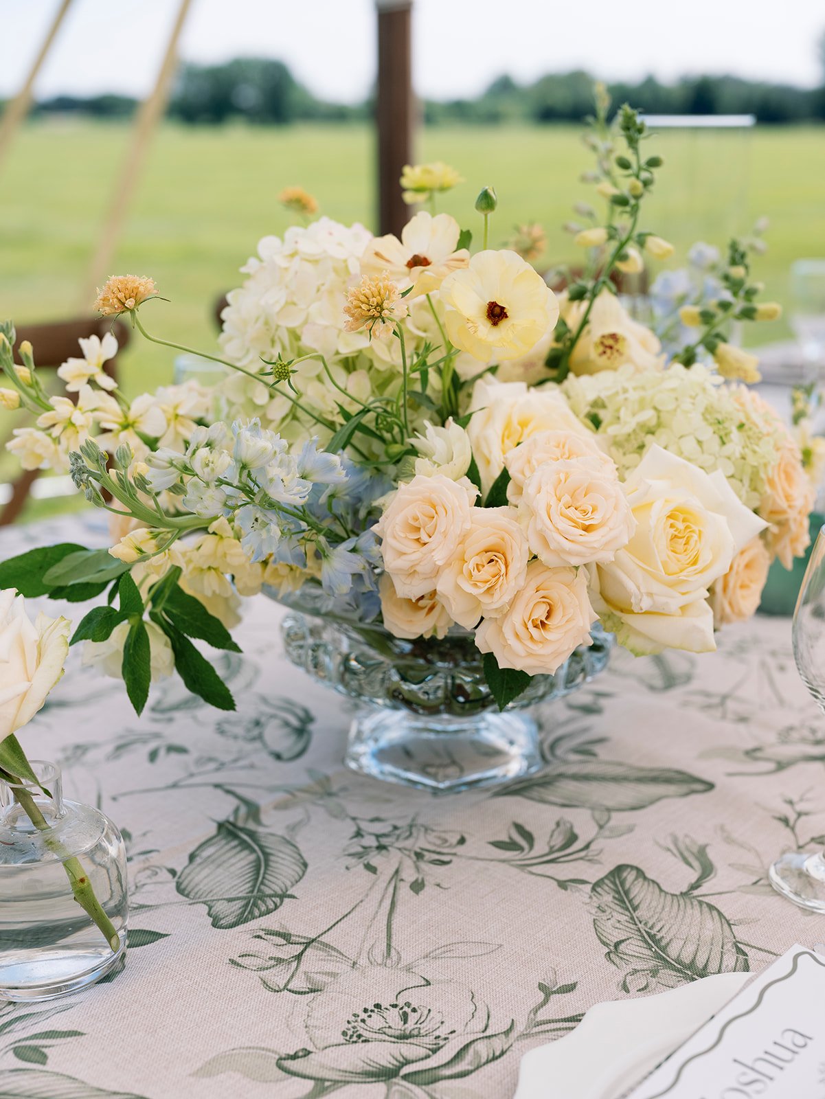 A floral arrangement with white and cream roses, hydrangeas, and other light-colored flowers in a glass bowl on a decorated table with leafy-patterned tablecloth, outdoors in a field setting.