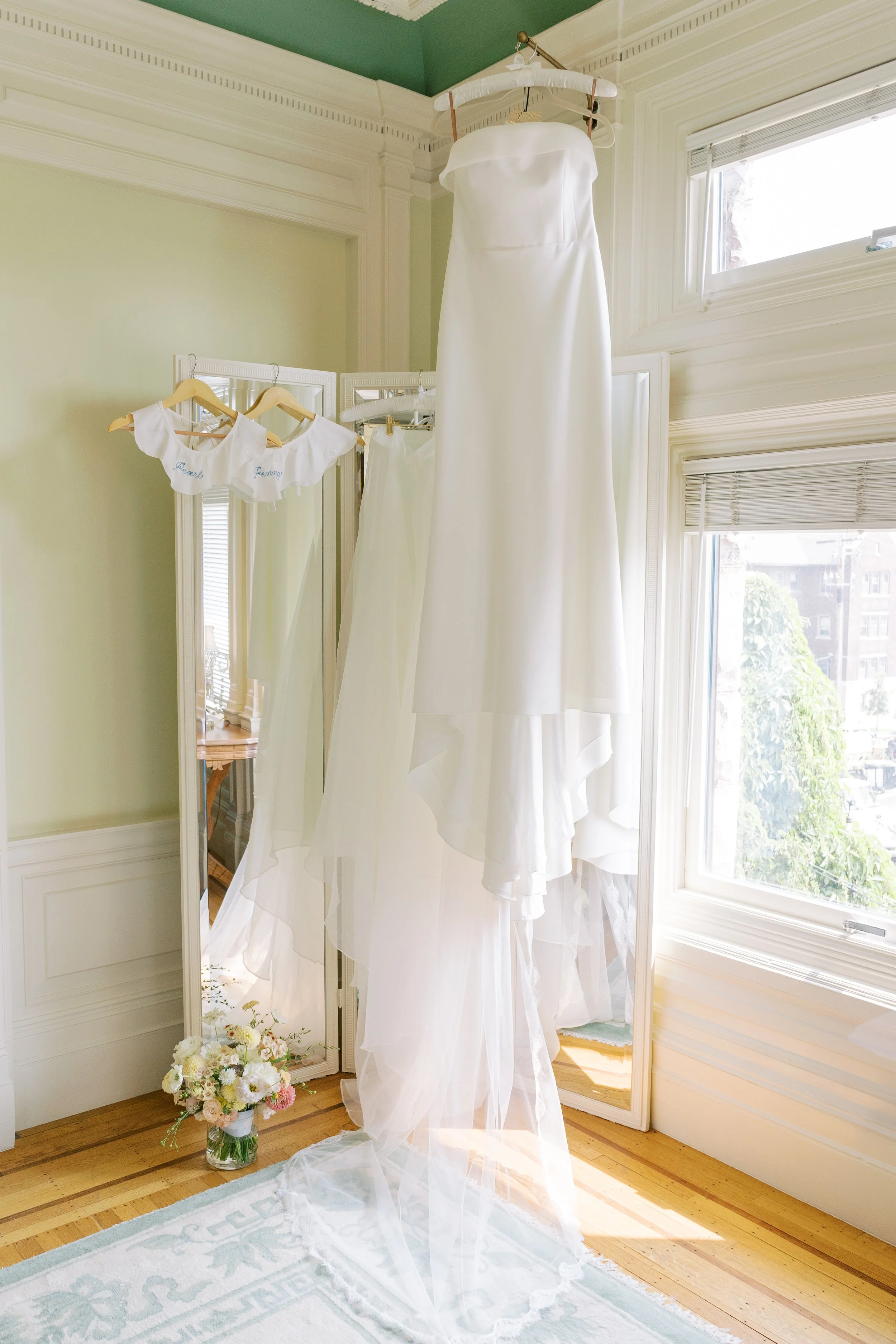 A white wedding dress hangs from a hanger, with a veil underneath, in a bright room near a window; two small white bodysuits with blue embroidery are hanging nearby, and a bouquet of flowers is on the floor.