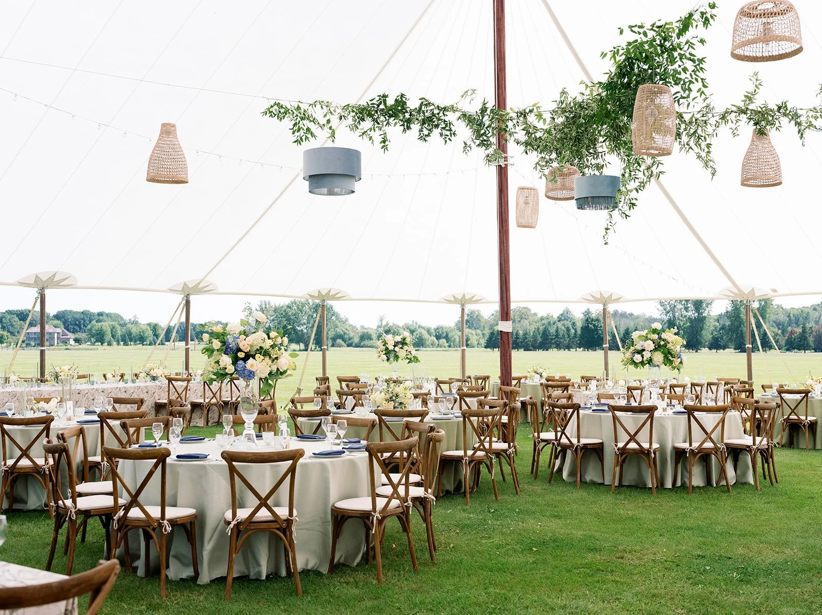 Overhead view of decorated outdoor event tent set up on grass, with round tables, floral centerpieces, dining ware, and wooden chairs. Decor includes hanging wicker and fabric lamps, with green foliage on tent ceiling, overlooking a scenic open field with trees in the distance.