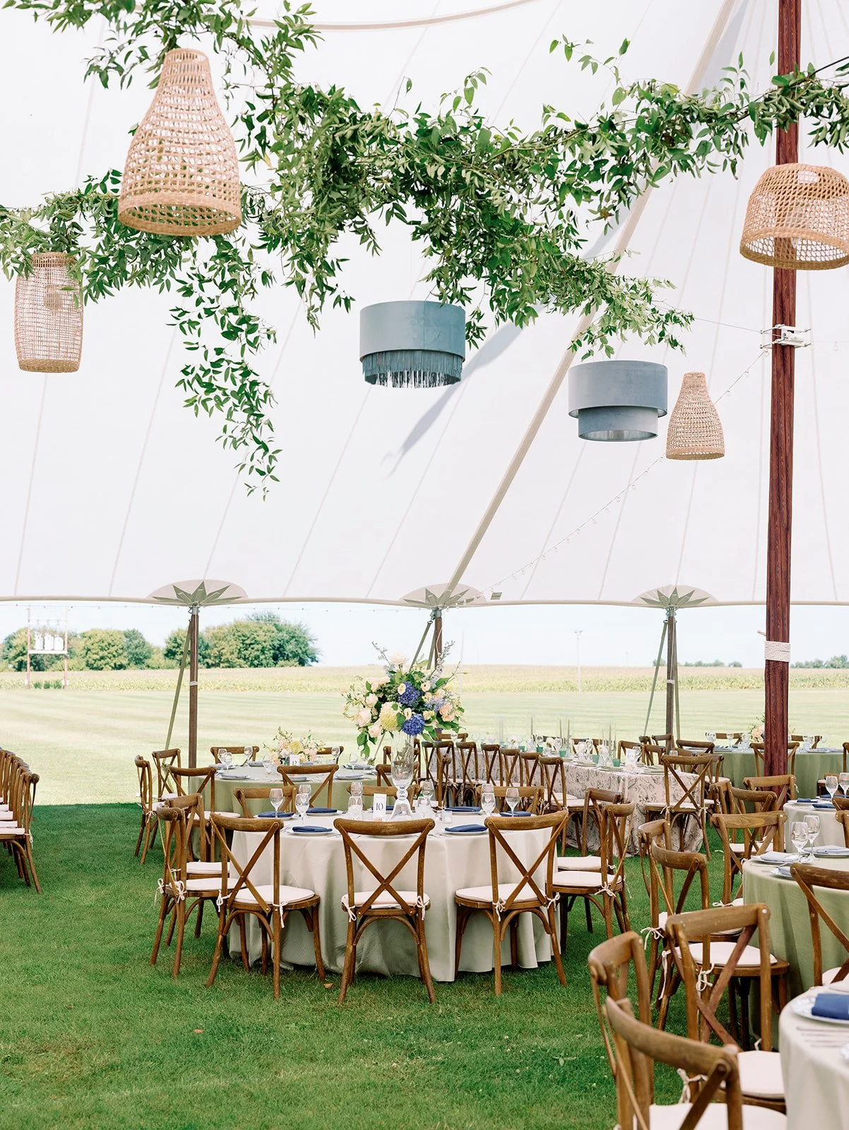 Outdoor wedding or event reception set up under a large white tent with round tables covered in white tablecloths, decorated with large floral centerpieces, surrounded by wooden chairs with white cushions, on a grassy field with open landscape in the background. Hanging from the tent ceiling are woven and fabric-covered pendant lights and greenery.