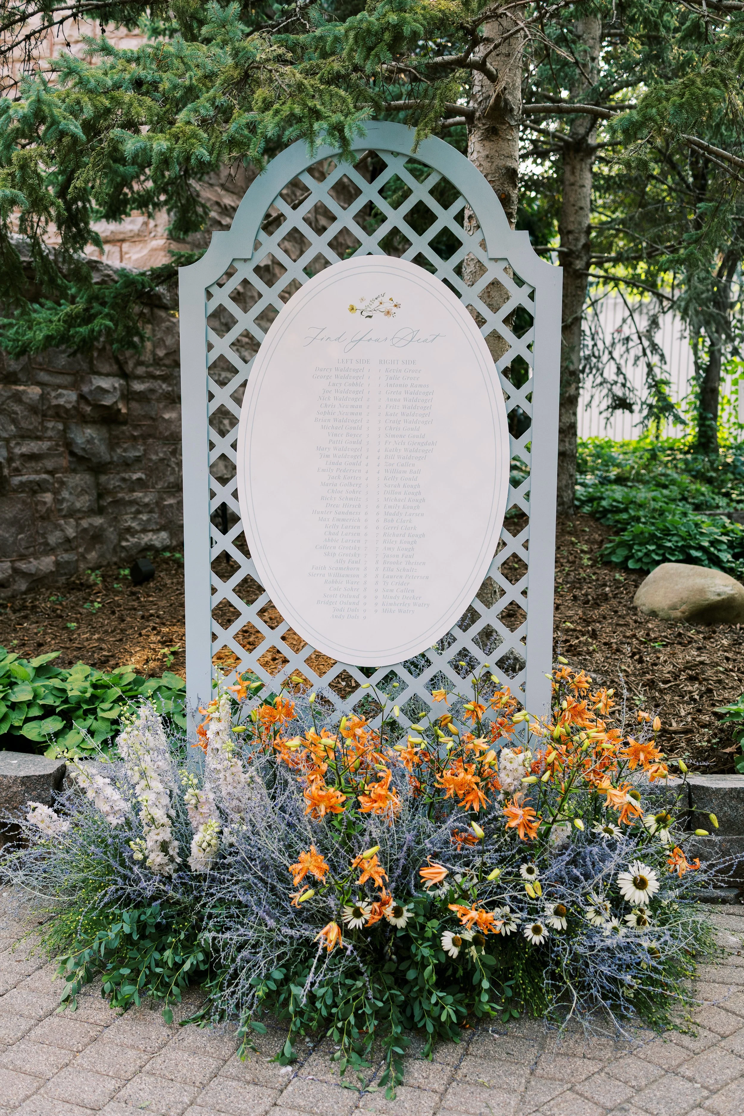 A memorial display featuring a large, white, oval-shaped list of names, set against a lattice frame with greenery and trees in the background. Bright orange and white flowers are arranged at the base.