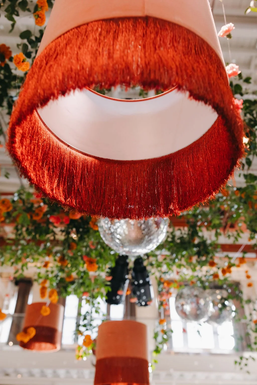Close-up of a hanging lampshade with red, fringed fabric, surrounded by floral decorations and other lighting fixtures in the background.