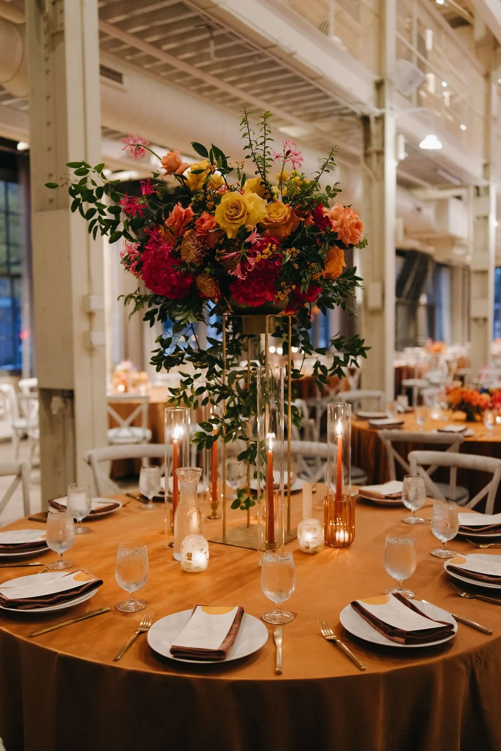 Elegant banquet table with a tall floral centerpiece of yellow, pink, and red flowers, surrounded by candles, glassware, napkins, and tableware in an industrial-style venue.