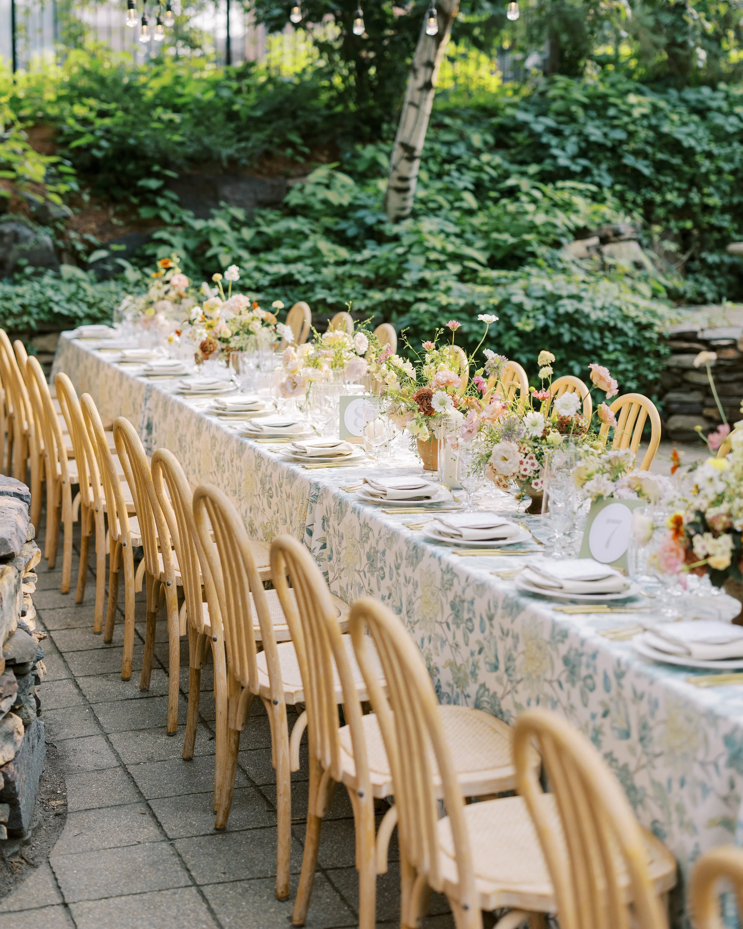 A long outdoor dining table set for a celebration, decorated with floral centerpieces, white plates, and glassware, surrounded by wooden chairs amidst lush green garden scenery.
