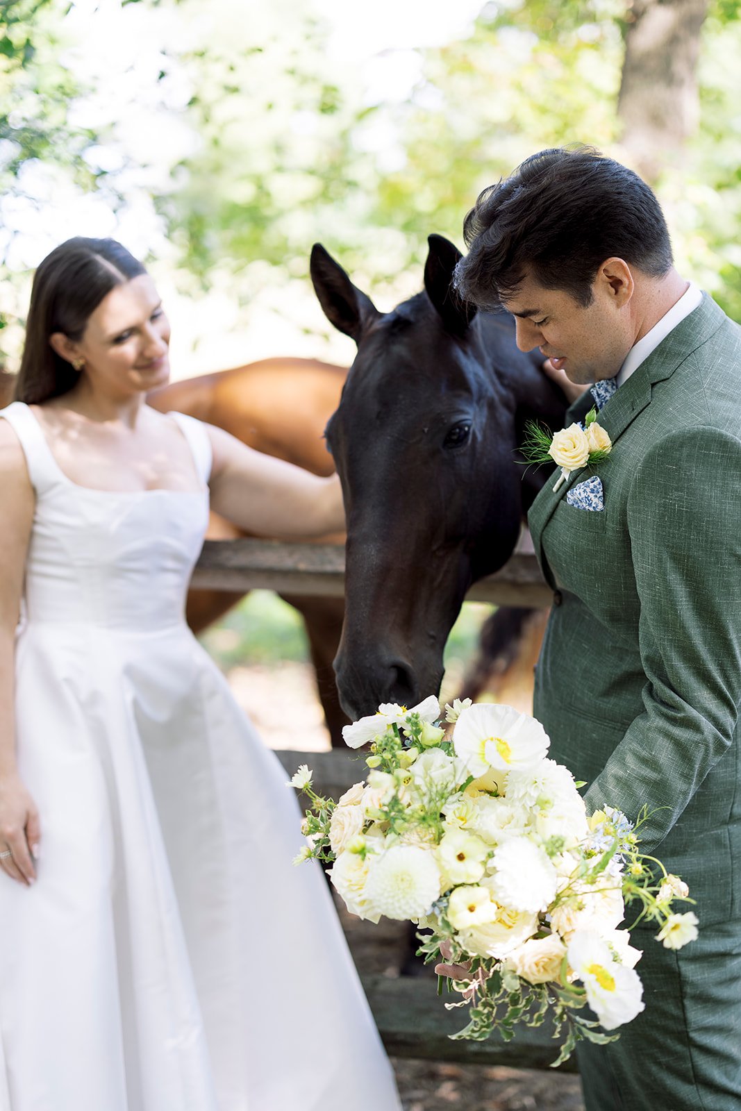 A bride and groom in wedding attire are outdoors, with the groom holding a large bouquet of white and yellow flowers. The groom is looking down and leaning his head gently against the face of a black horse, while the bride, dressed in a white gown, is smiling and gently touching the horse's back. The background features green foliage and a wooden fence.