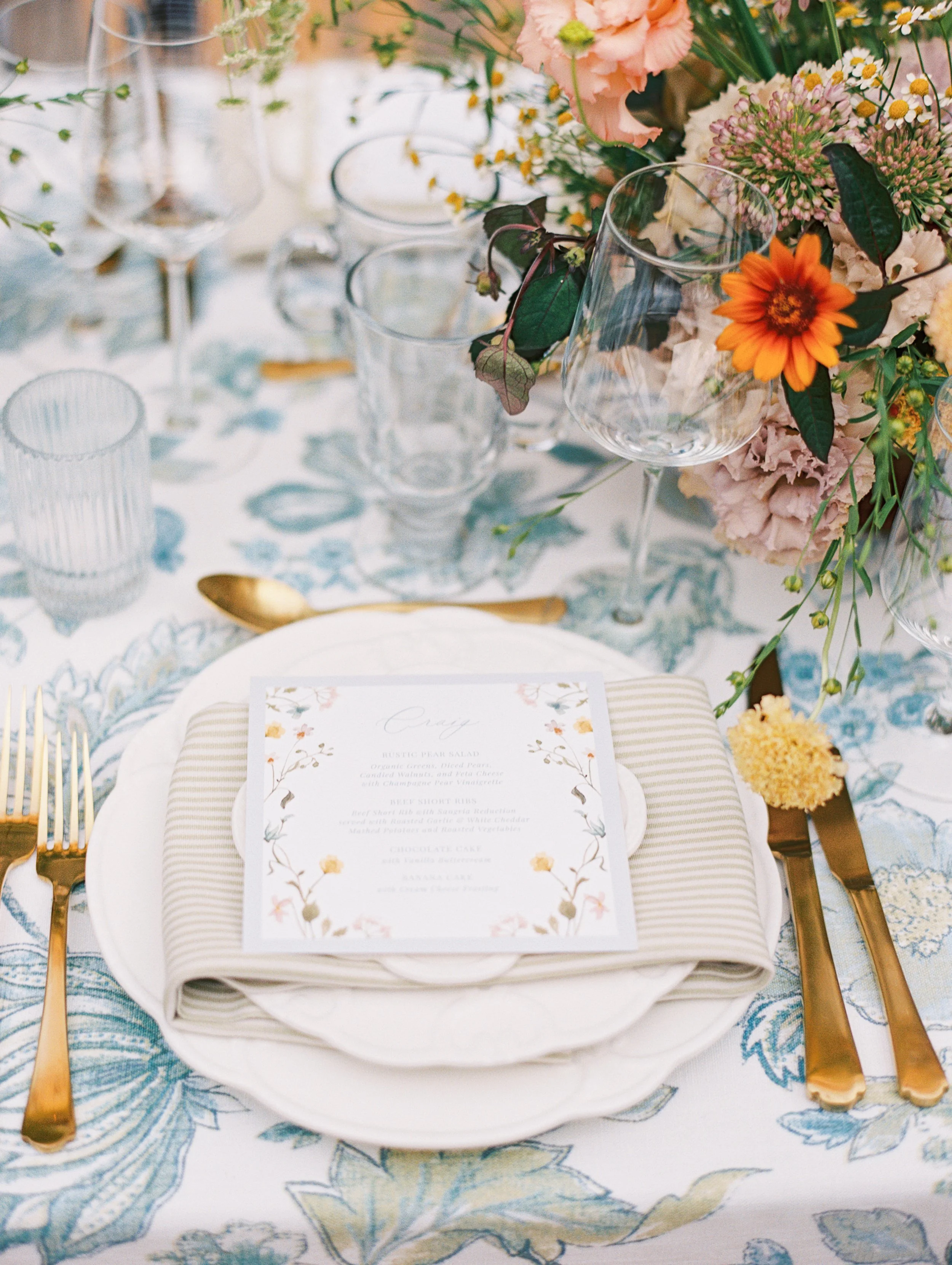 A table setting with a white plate, gold utensils, a floral menu card, and a floral centerpiece of pink, orange, and cream flowers with greenery in a glass vase.