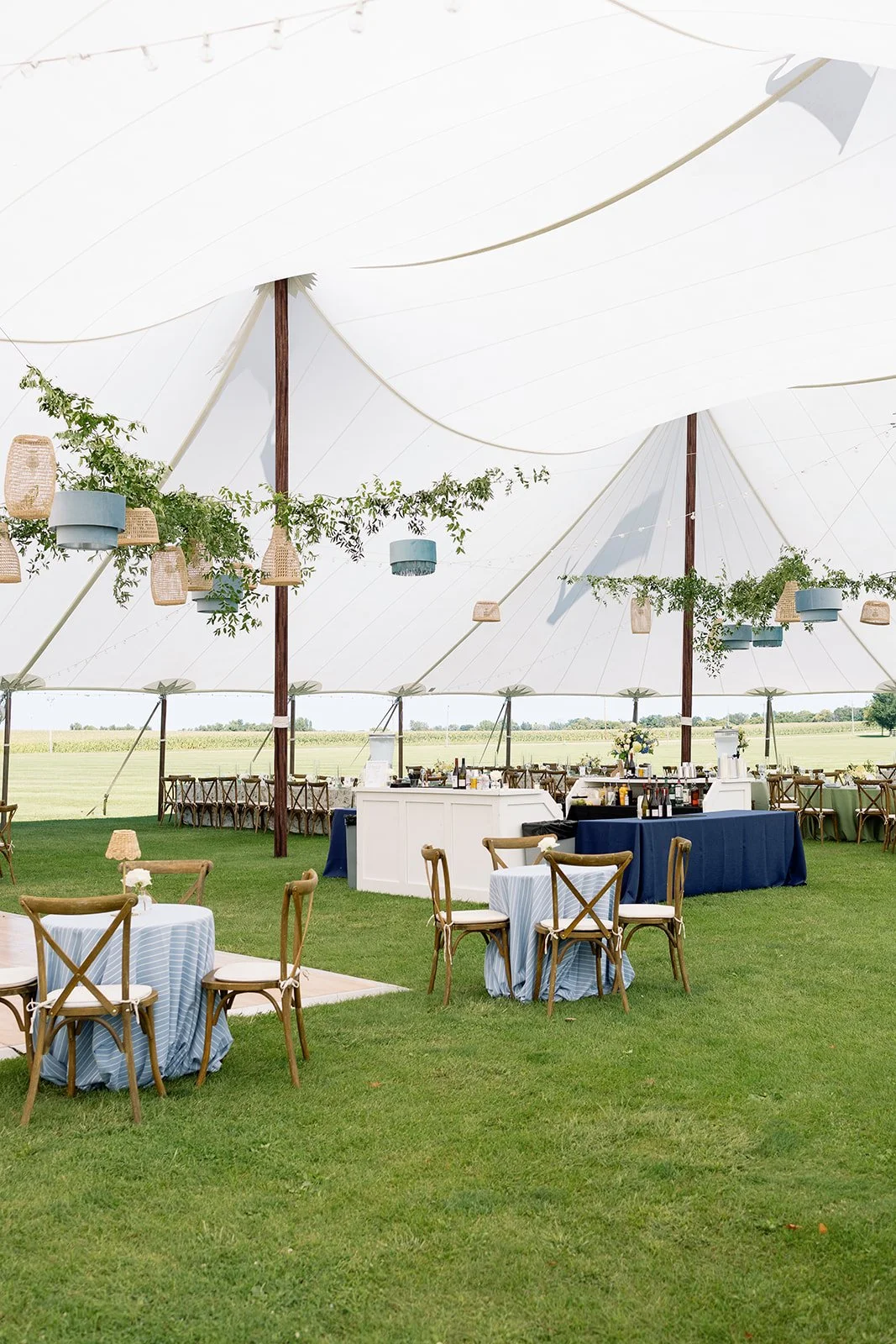 Outdoor wedding or event setup under a large white tent with tables and chairs and hanging lanterns and greenery decor.
