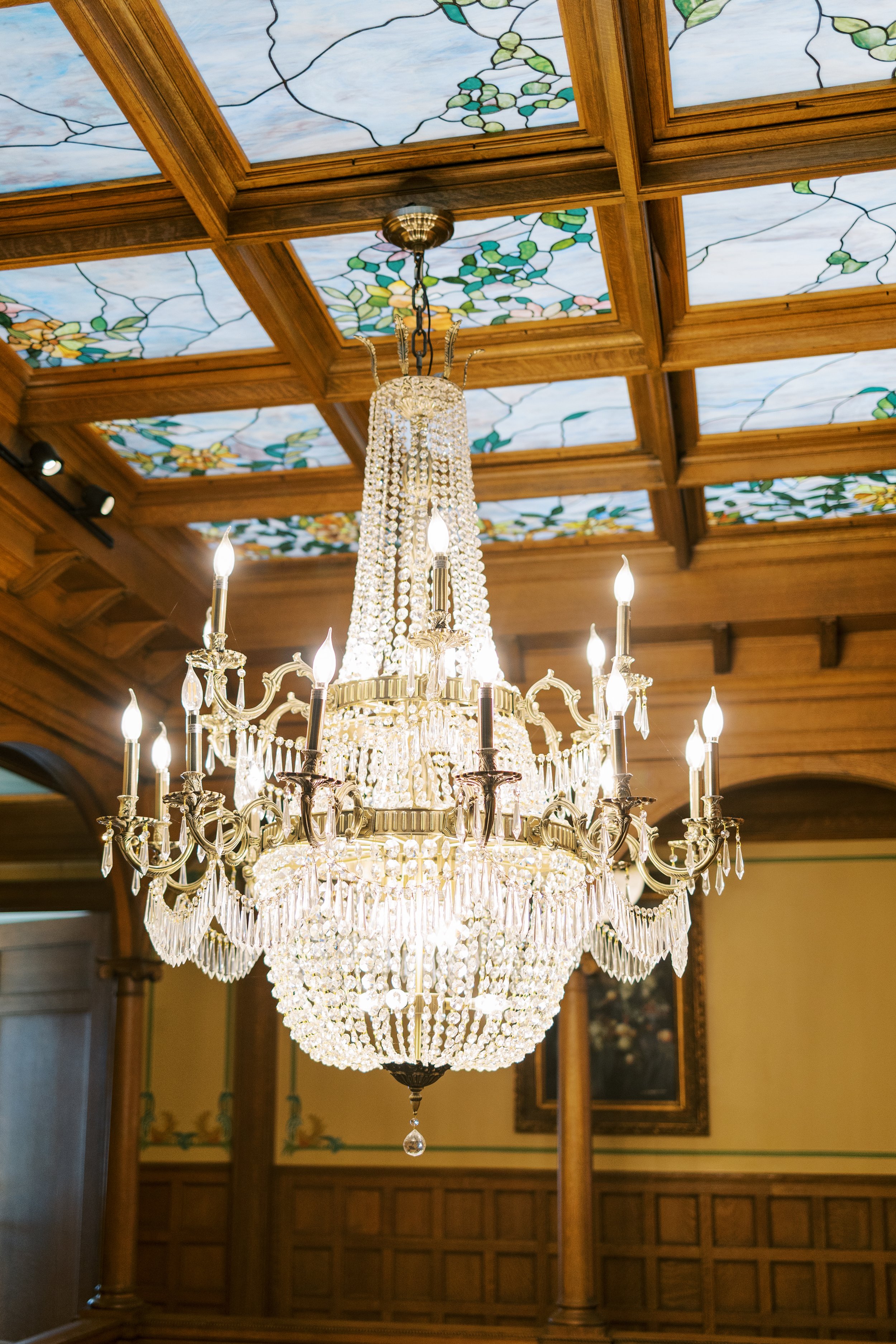 Large ornate chandelier with crystal embellishments hanging from a wooden coffered ceiling with stained glass panels.
