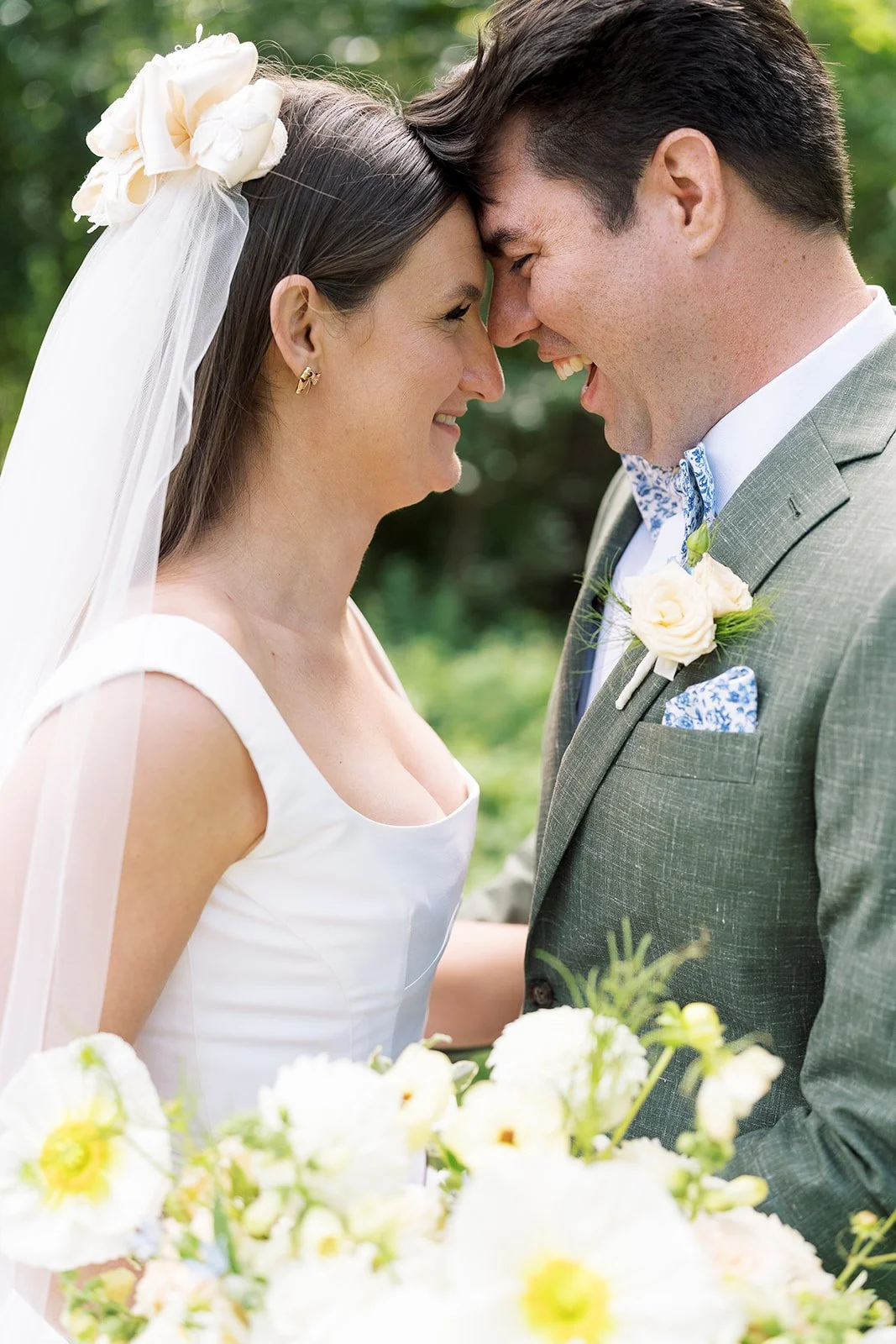 A bride and groom with foreheads touching, smiling at each other outdoors with greenery in the background, during their wedding.