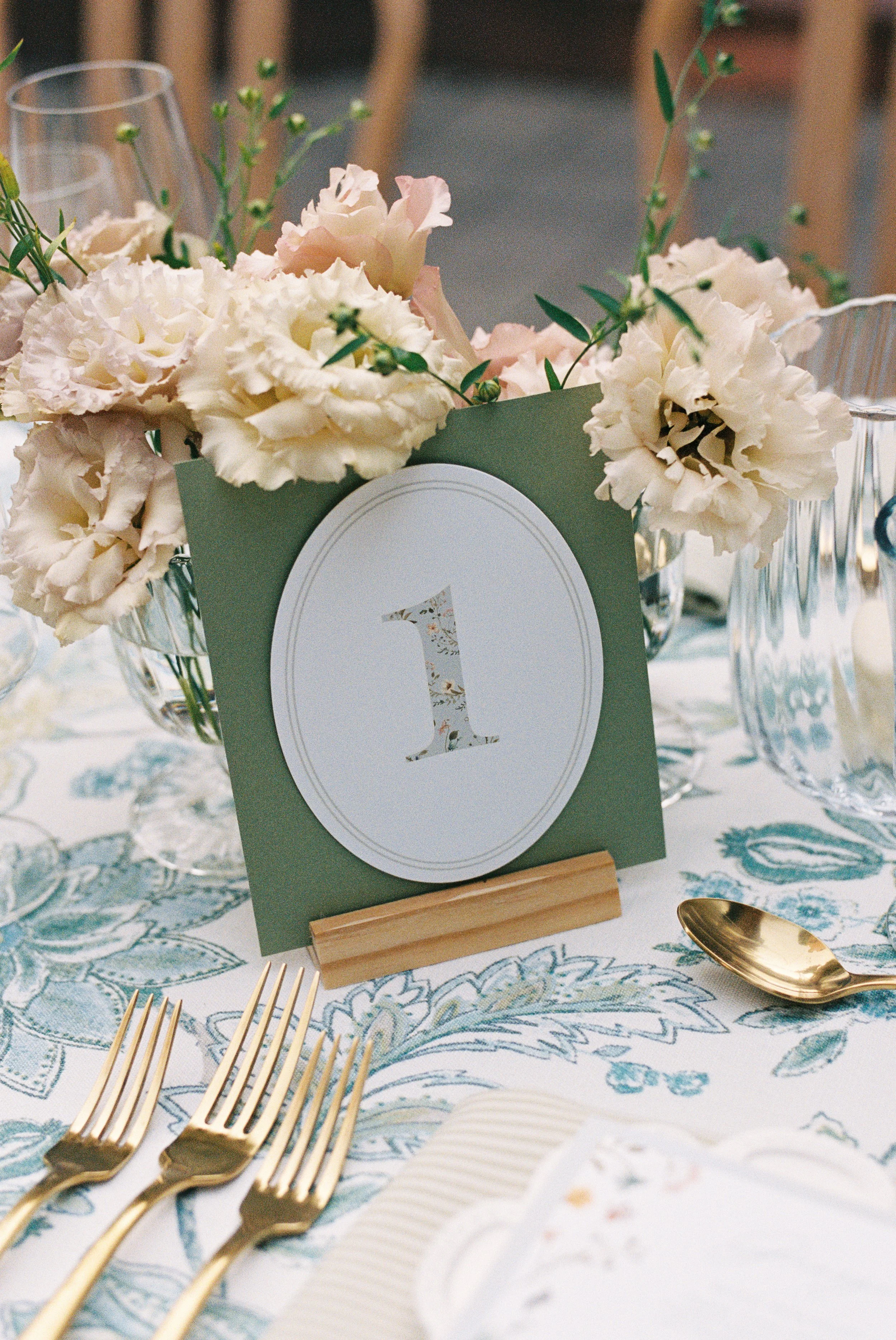 Table setting with gold utensils, a floral centerpiece with cream and blush flowers, and a table number card with the number one on a wooden stand.