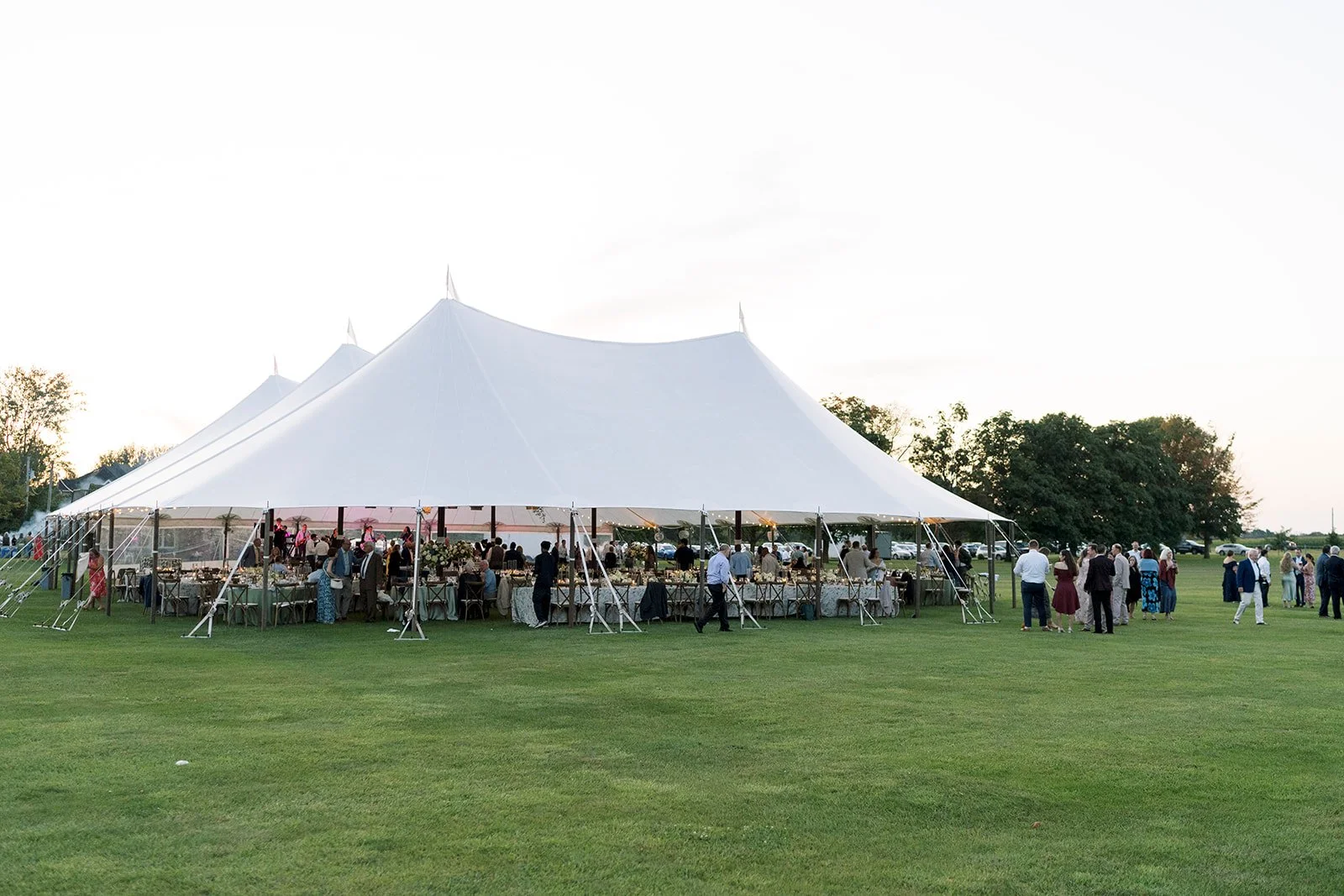 Large outdoor event tent set up on a grassy field with people gathered underneath and outside, celebrating during late afternoon or early evening.