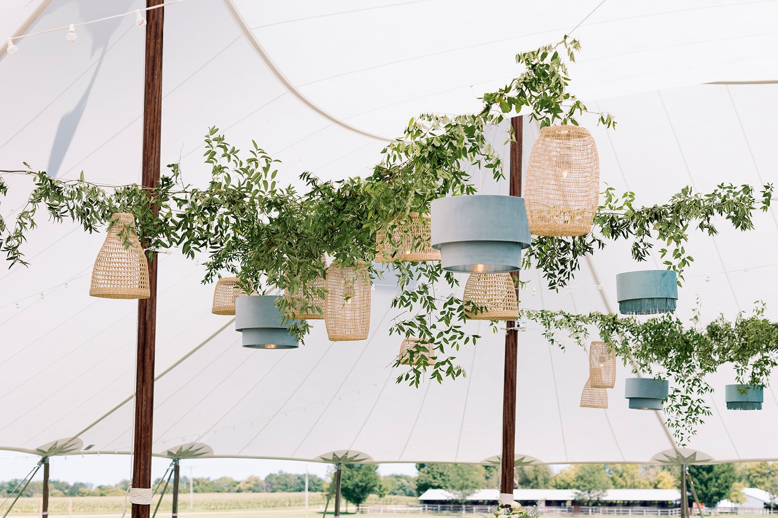 Hanging wicker and blue fabric lanterns with greenery under a white tent at an outdoor event.