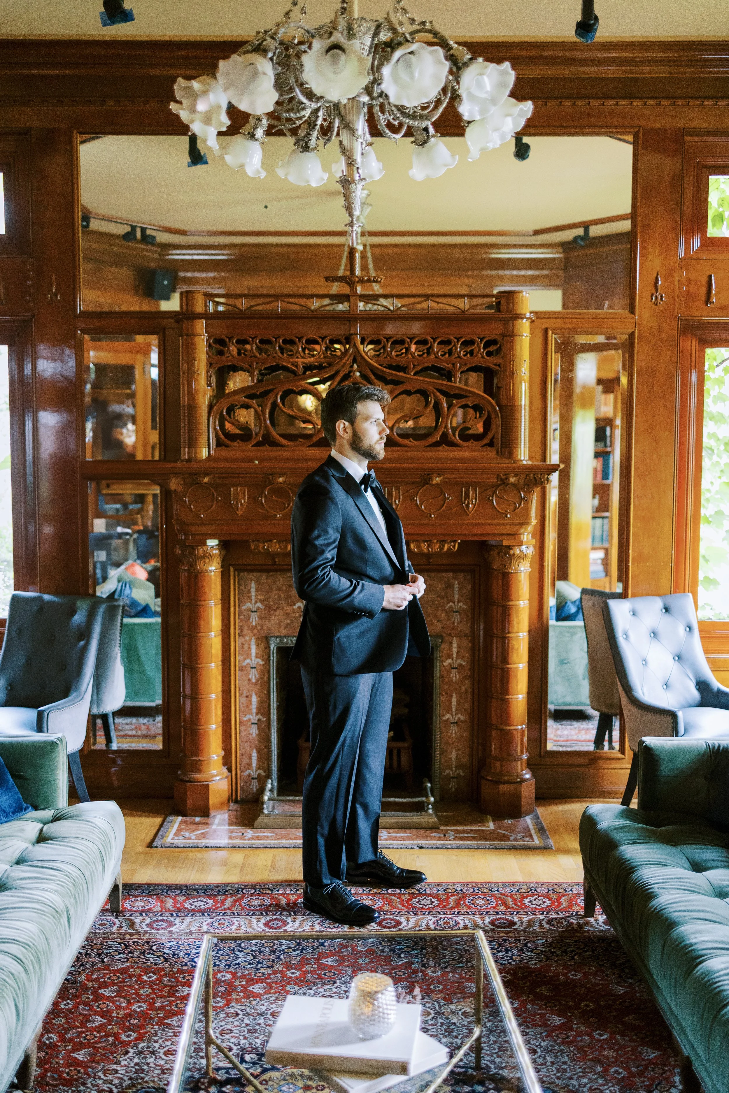 A man in a black tuxedo standing in a luxurious wood-paneled room with ornate fireplace, large mirror, and plush furniture; he adjusts his cuffs, preparing for a formal event.