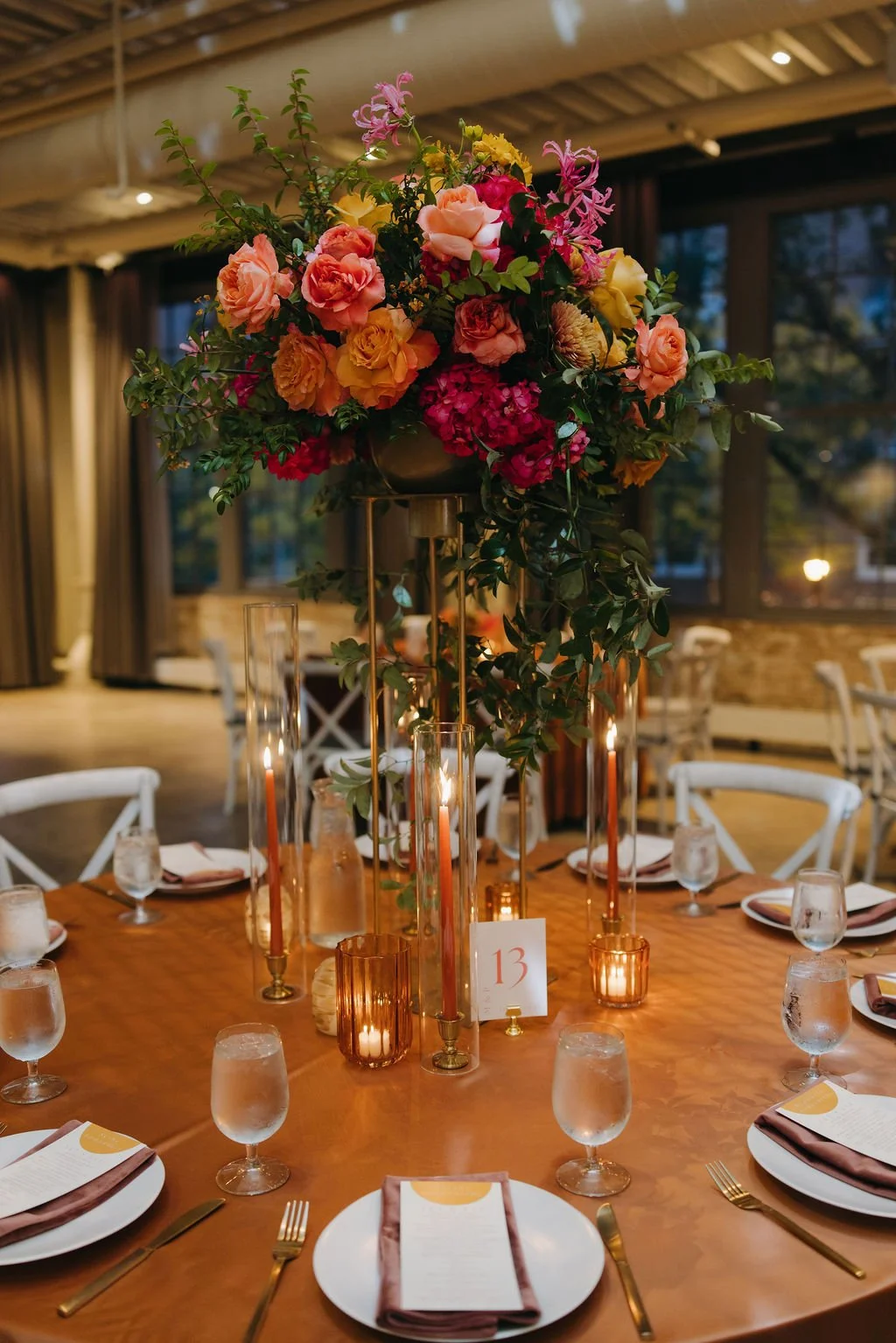 A decorated round table with a tall floral centerpiece of pink, red, yellow, and peach roses, and green foliage. The table has candles in glass holders, water glasses, and place settings with menus and napkins. There is a small table number card with the number 13. The background shows large windows, curtains, and a warmly lit interior.