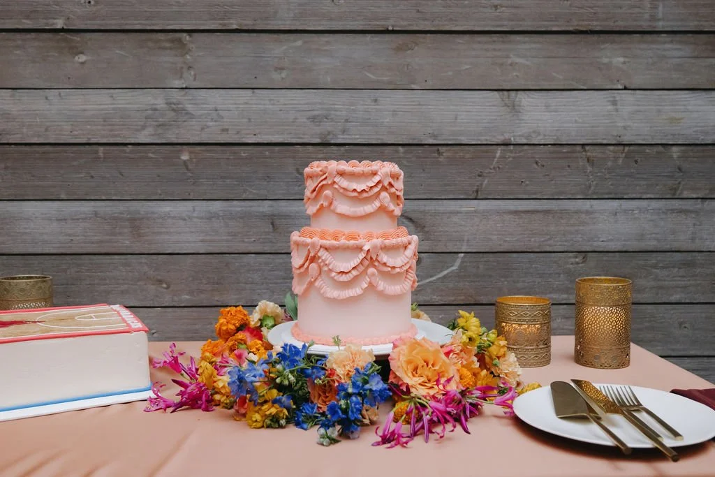 A pink two-tiered cake decorated with pink frosting ruffles, surrounded by colorful flowers, on a table with gold candle holders, plates, cutlery, and a large book, against a wooden fence background.