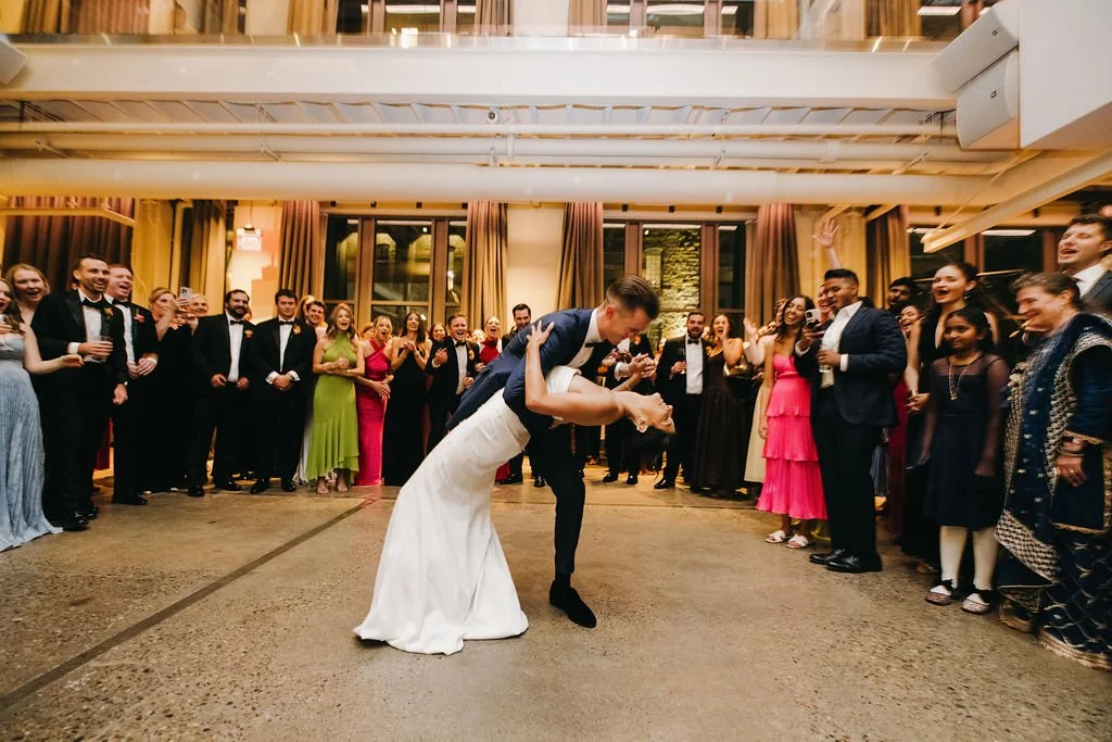 A bride and groom are dancing closely in the center of a large group of guests at a wedding reception, with everyone watching and smiling.