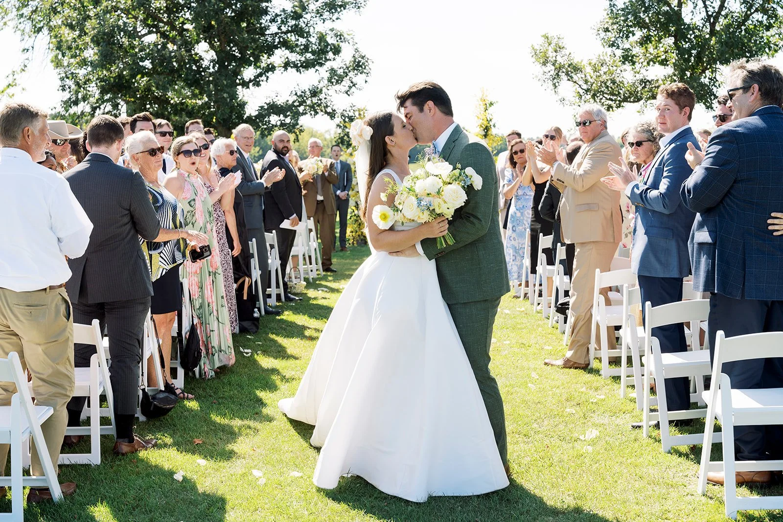 A newlywed couple sharing a kiss during their outdoor wedding ceremony, surrounded by seated and standing guests clapping and taking photos on a sunny day with green trees in the background.