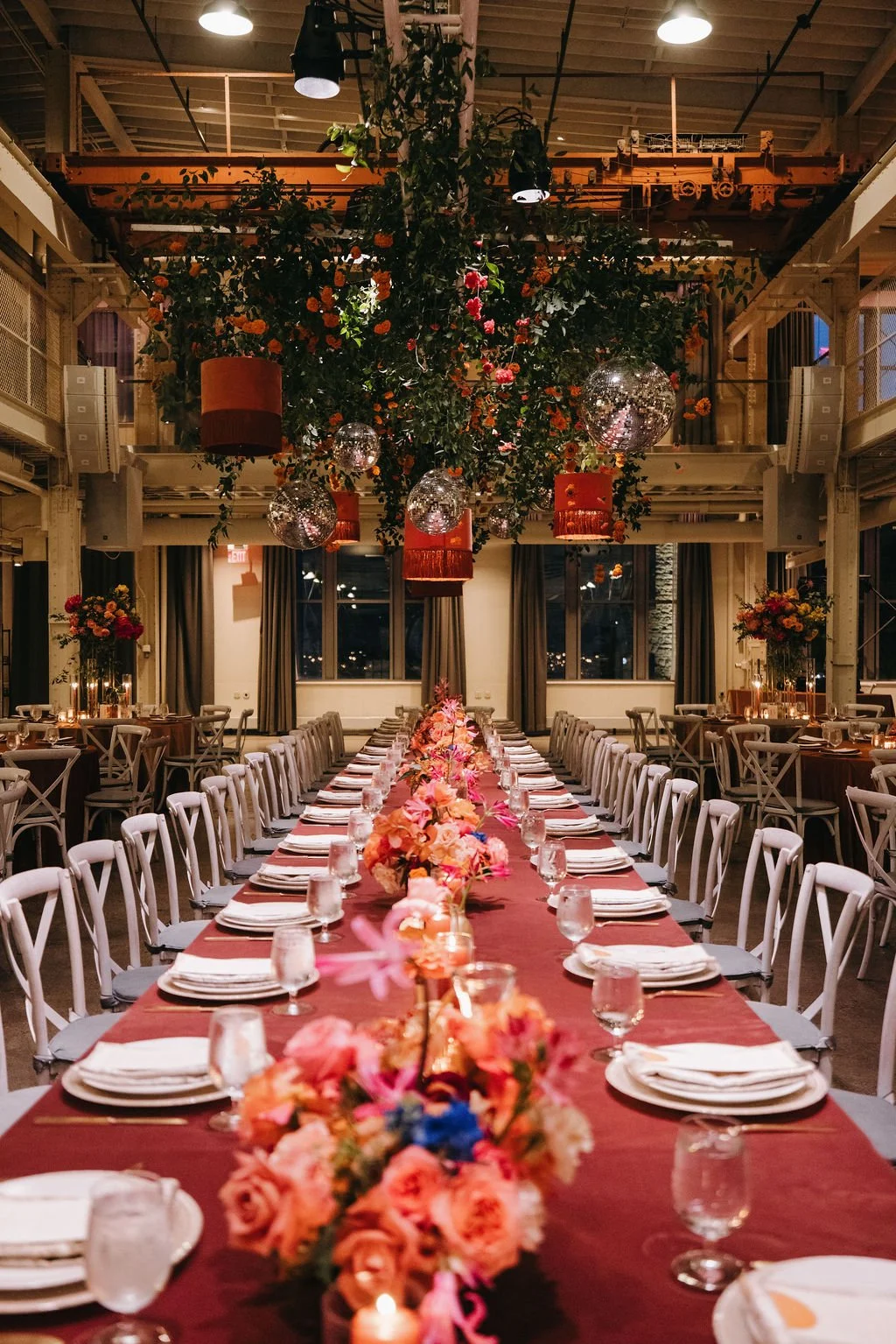 A long banquet table set for a celebration with pink and peach floral arrangements, surrounded by white chairs, in a decorated event space with hanging floral and disco ball decorations.