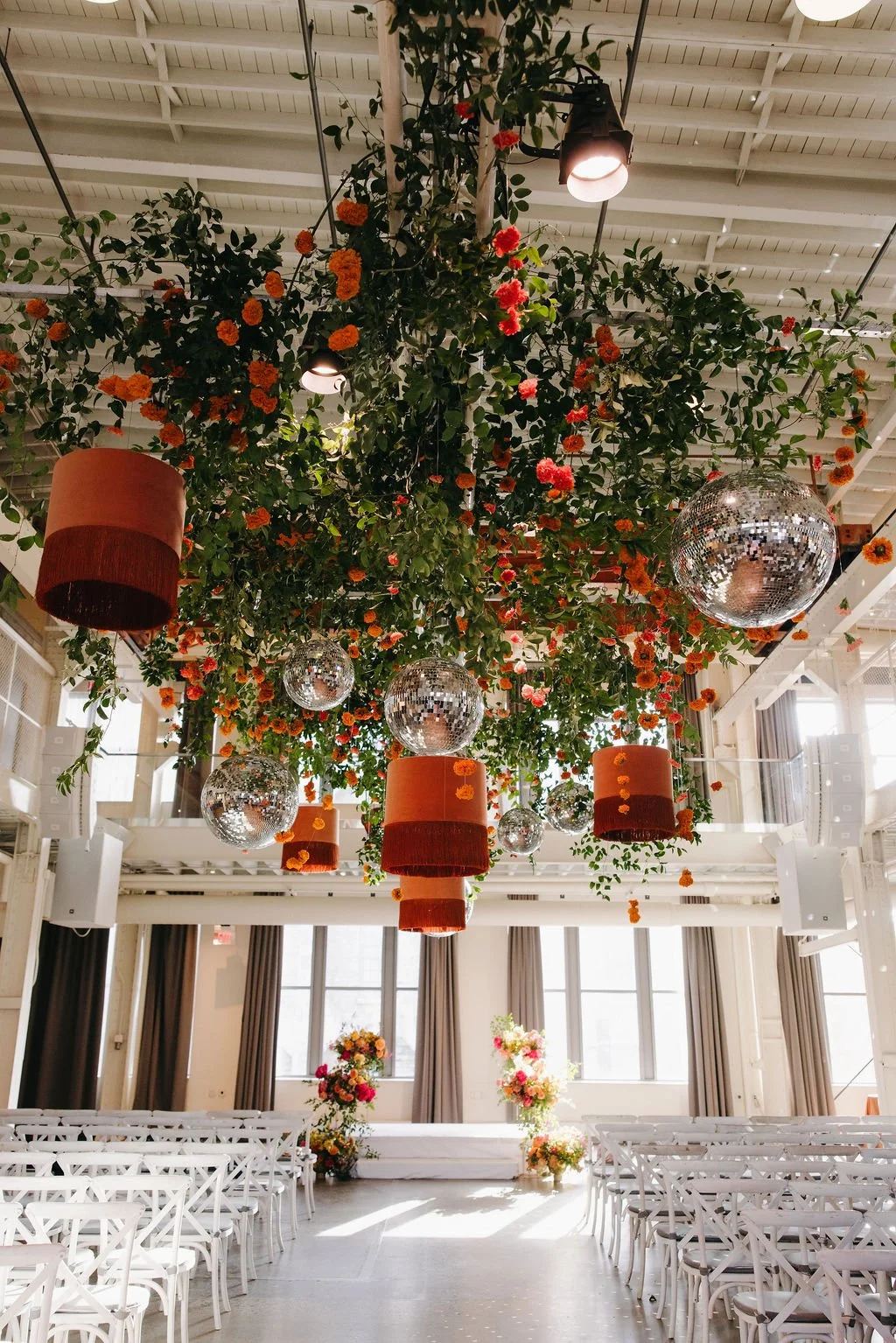 Indoor event space decorated with hanging greenery, orange flowers, disco balls, and hanging lamps with shades, set up with white chairs and floral arrangements in the background.