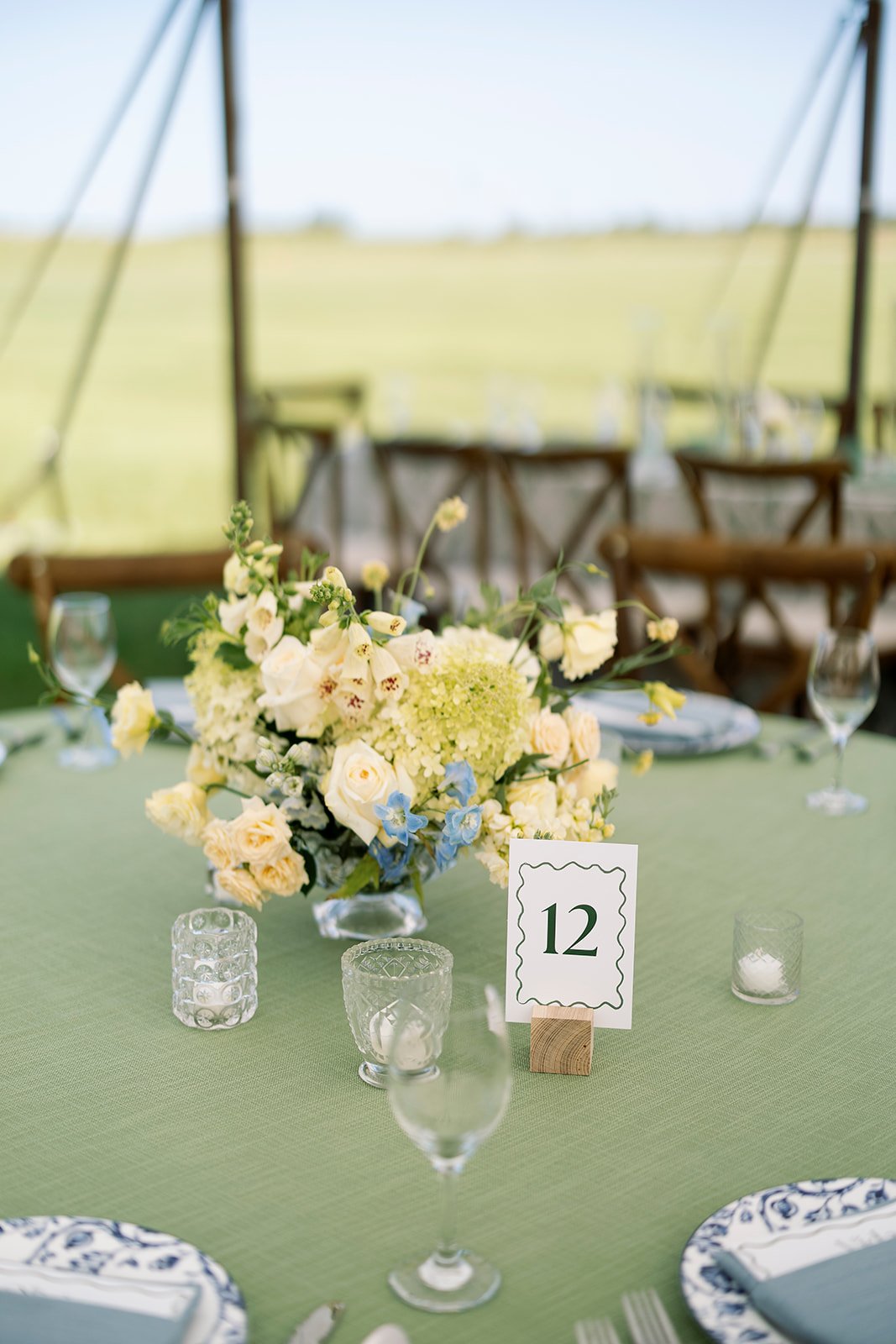 Table setting with a floral centerpiece on a green tablecloth, set for a wedding or event, with a table number 12 sign, wine glasses, and candles, outdoors with chairs and a grassy field in the background.