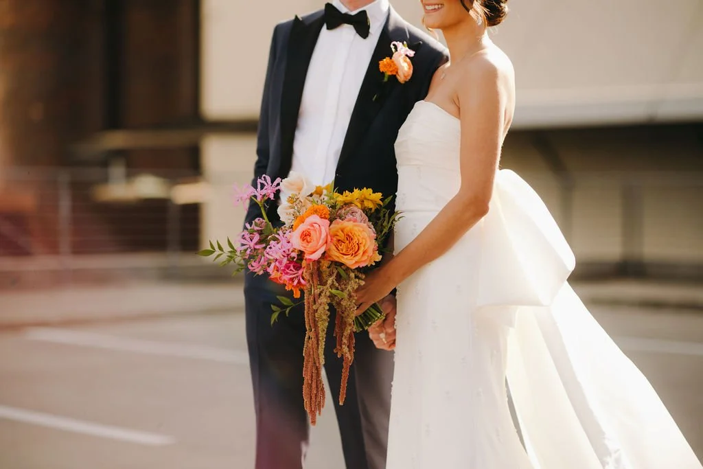 Close-up of a bride and groom holding a floral bouquet at their wedding, with the bride in a white strapless wedding gown and the groom in a tuxedo with a black bow tie.